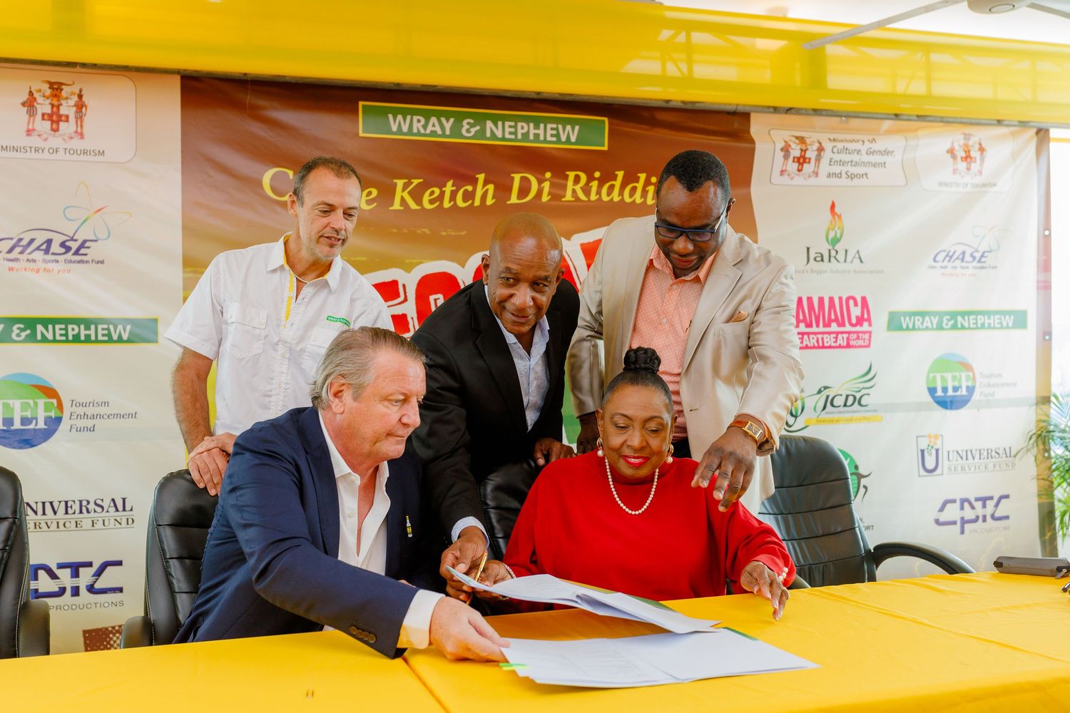Officials signing documents at a Wray & Nephew Catch Di Riddim event, with sponsors and partners logos displayed behind them.