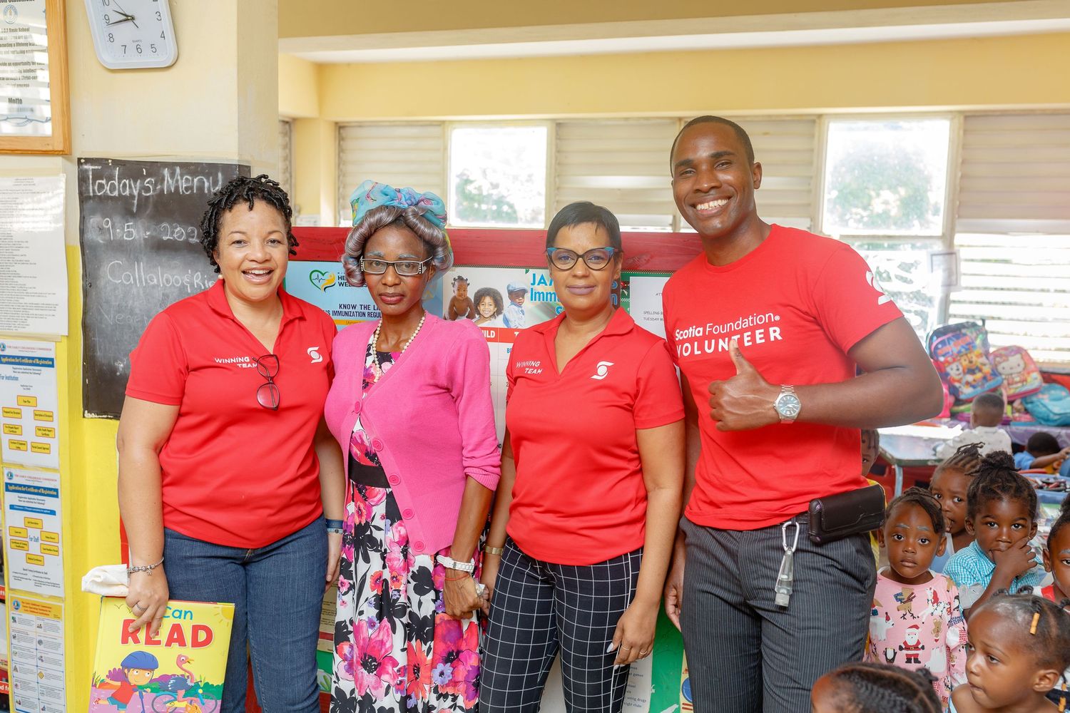 Four volunteers in red shirts pose together in a colorful classroom, smiling for a group photo.