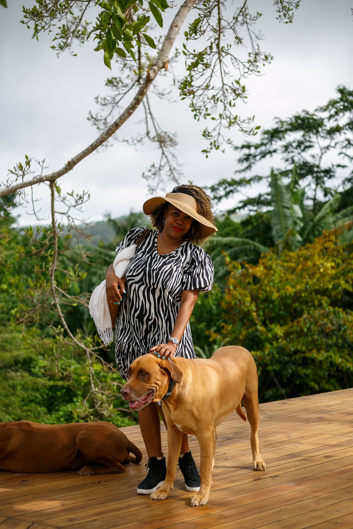 Woman in zebra dress and sun hat petting a tan dog on a wooden deck surrounded by lush tropical greenery.