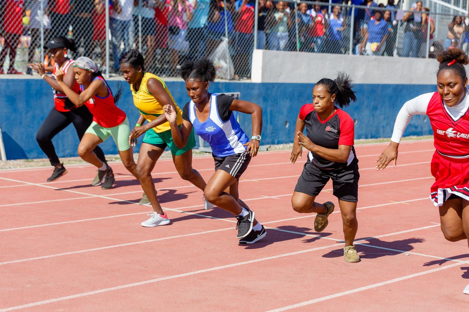 Female track athletes sprint from starting blocks during a competitive race on a blue outdoor running track.