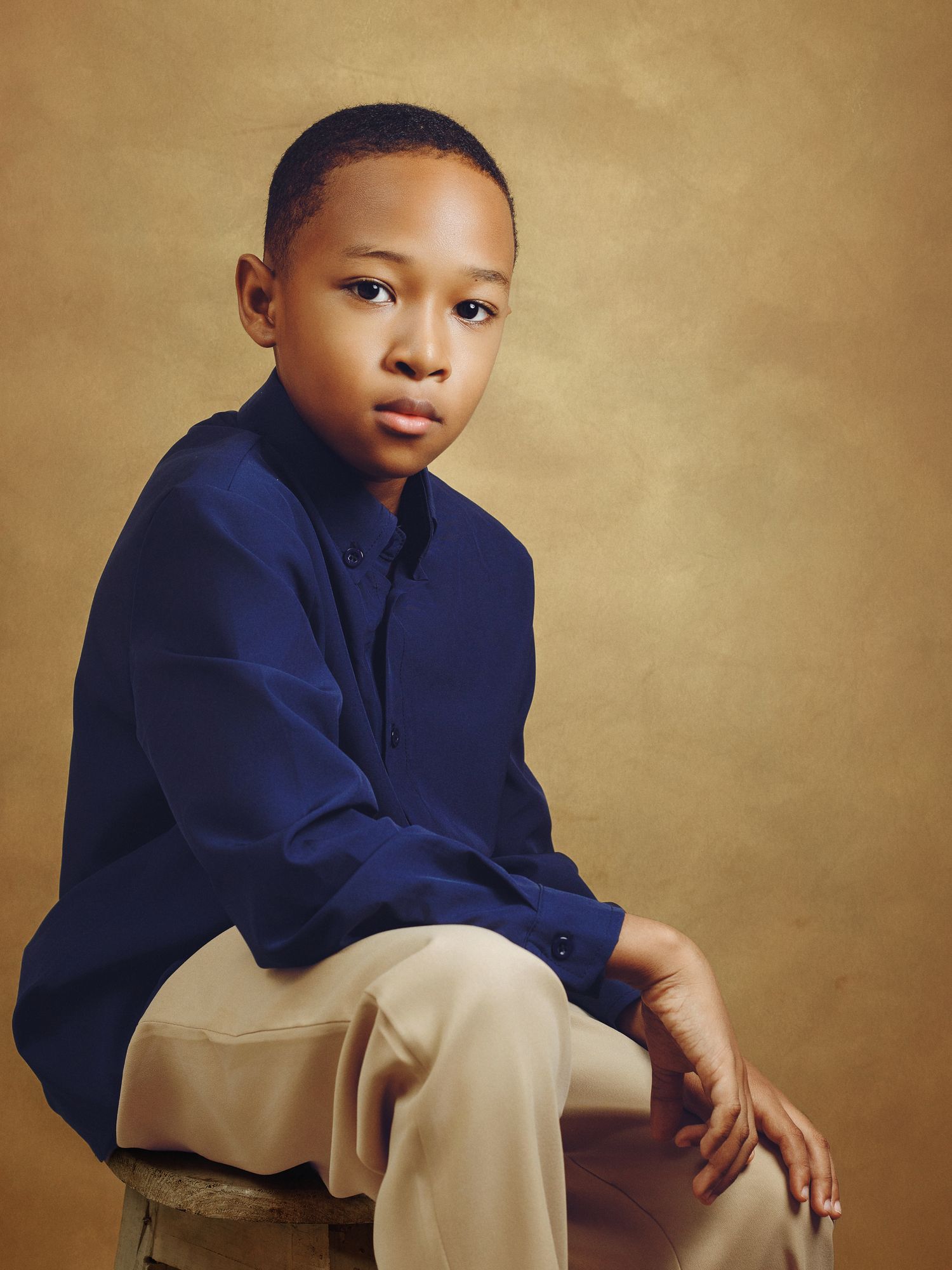 Young child in navy blue shirt and khaki pants poses against tan backdrop in professional portrait series.