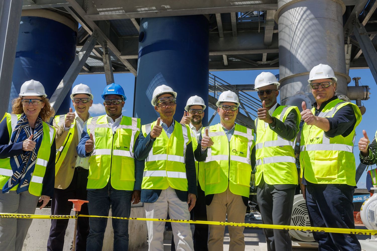 Group of workers in yellow safety vests and hard hats giving thumbs up at an industrial facility.
