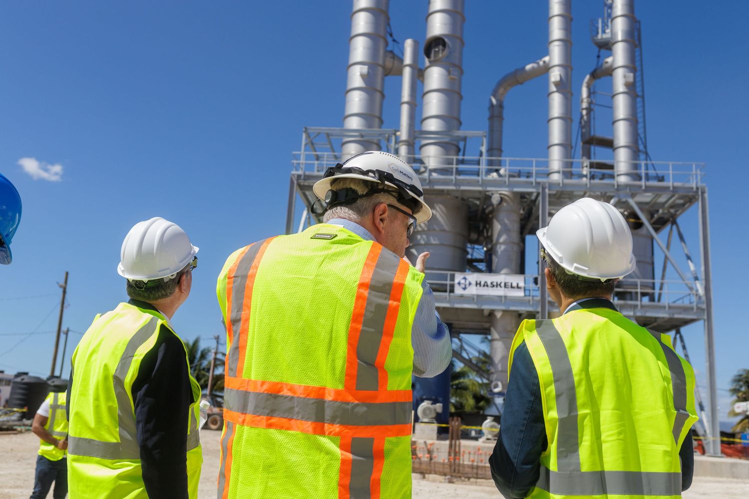 Three engineers in safety vests and hard hats inspect an industrial facility with tall metal stacks under blue sky.