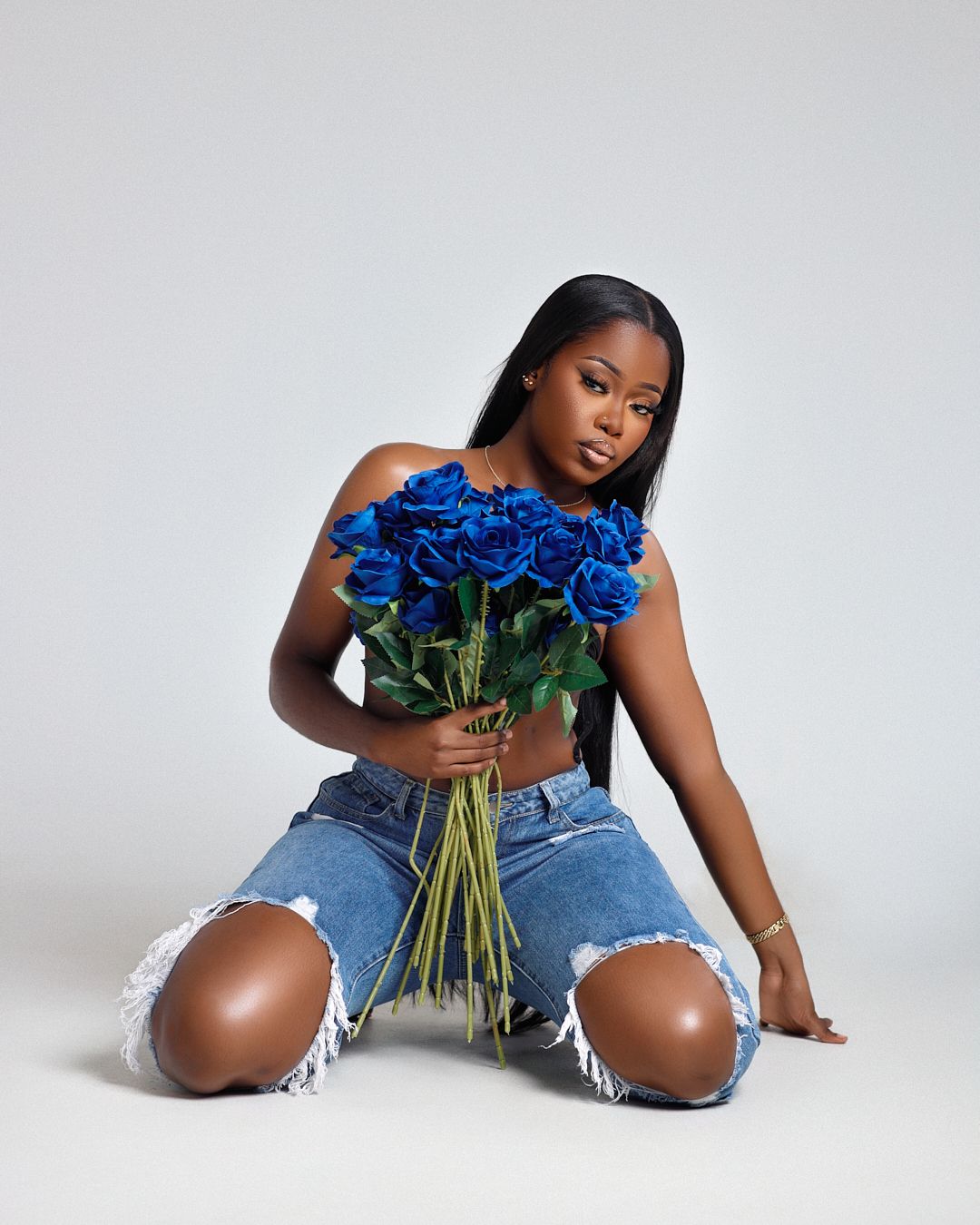 Model in ripped jeans poses with blue roses against white studio background.