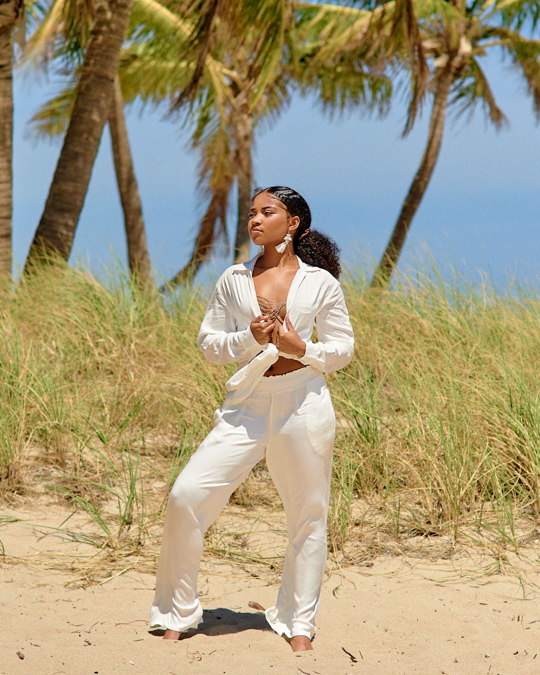 Model in all-white outfit poses on sandy beach with palm trees.