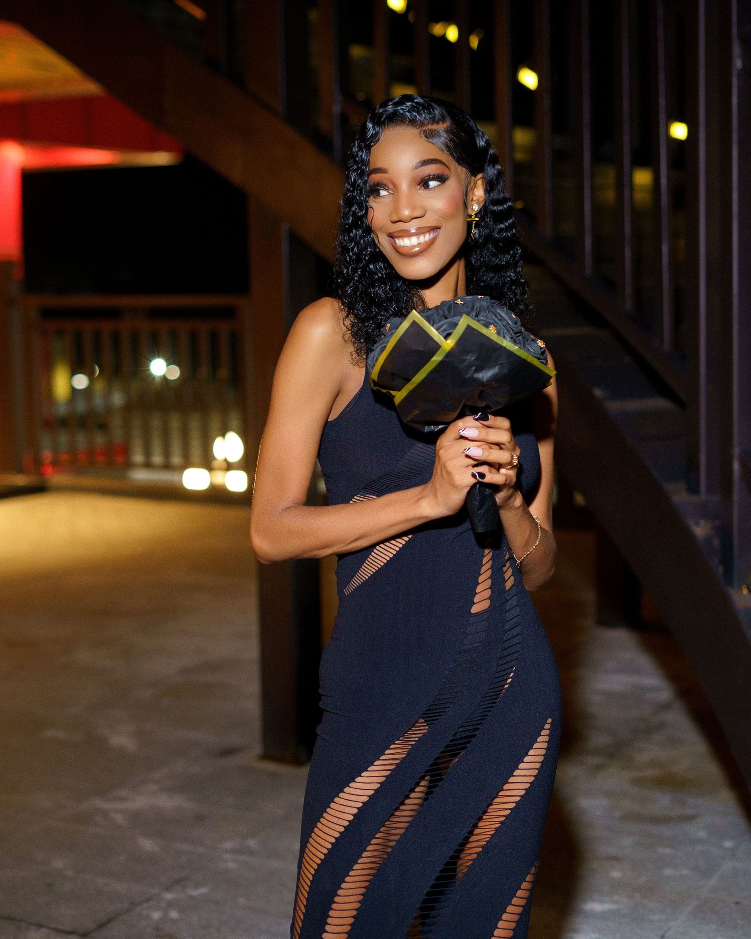 A woman in a black dress poses confidently at night with green lanterns glowing in the background.