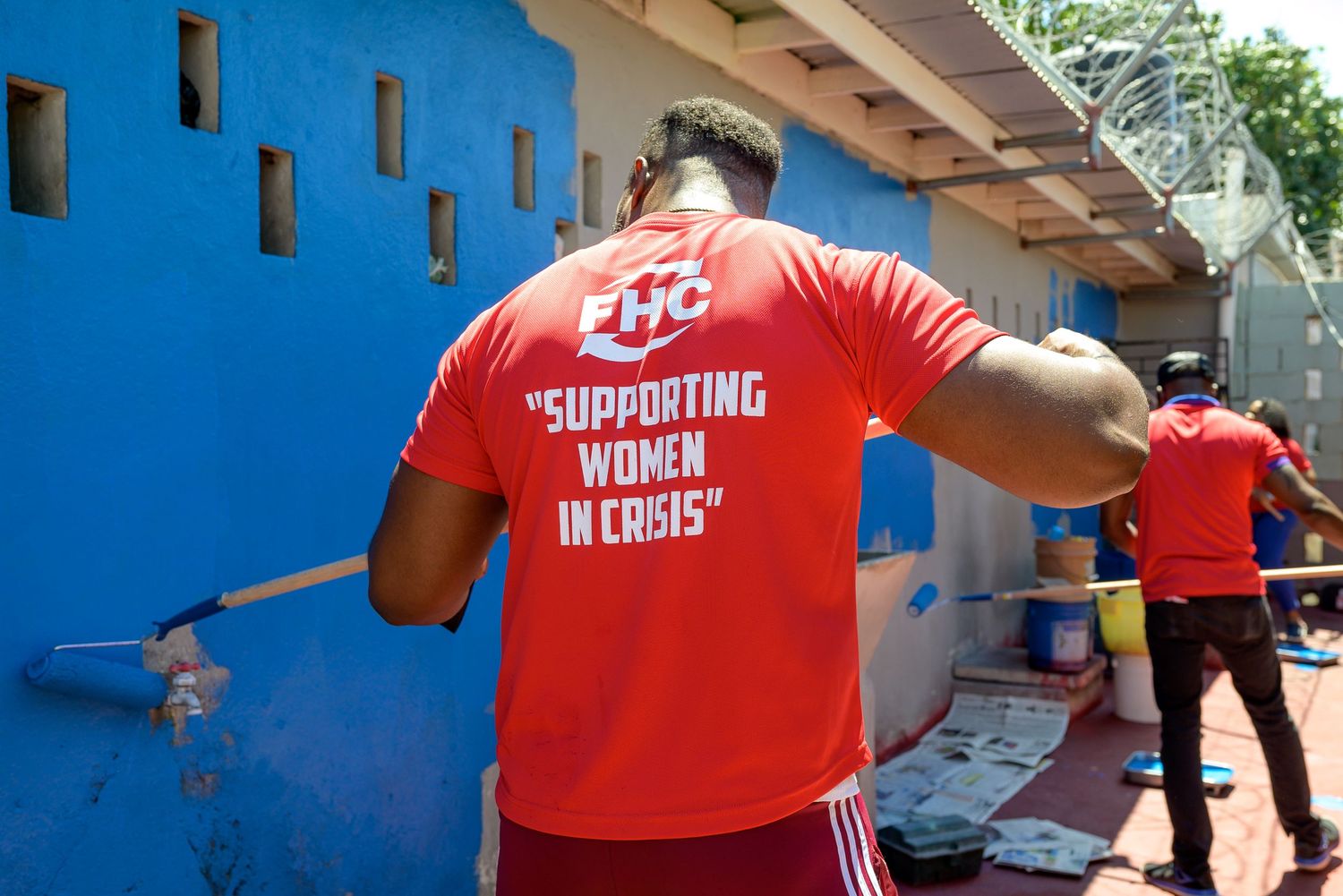 Volunteers in red FHC shirts supporting women in crisis paint a blue wall outdoors.