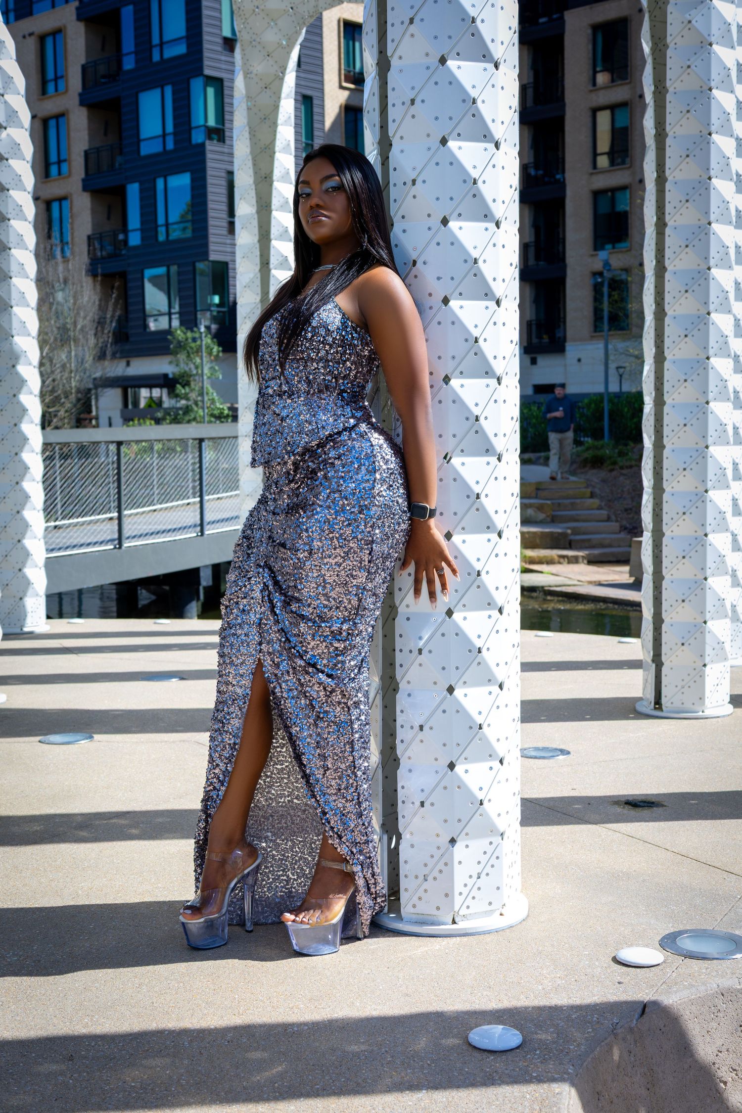 Woman in silver sequin slit gown posing beside white lattice columns in urban setting.