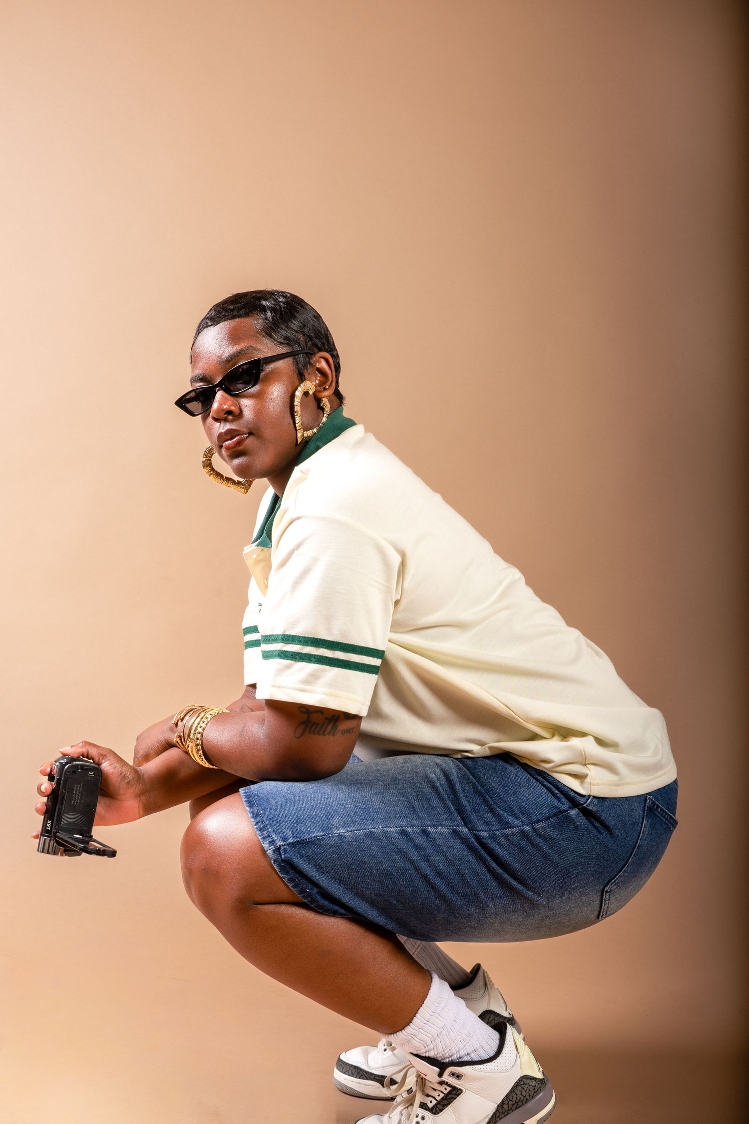 Person crouching in denim shorts, yellow polo, sunglasses, gold jewelry, and sneakers against a beige background.
