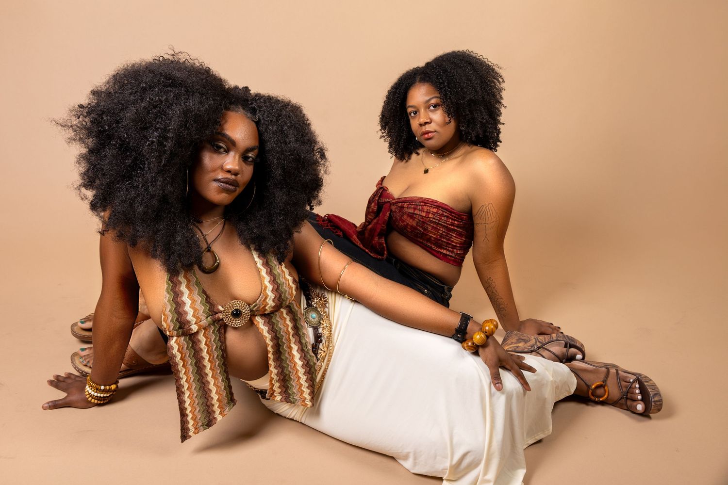 Two Black women with natural afro hair pose confidently in a beige studio, wearing stylish crop tops and wide-leg pants.