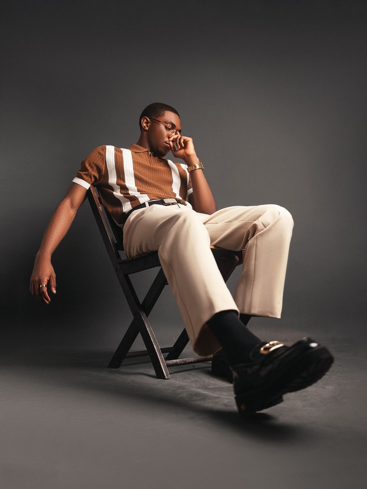 Stylish man in striped brown polo and cream trousers relaxing on a folding chair in studio.