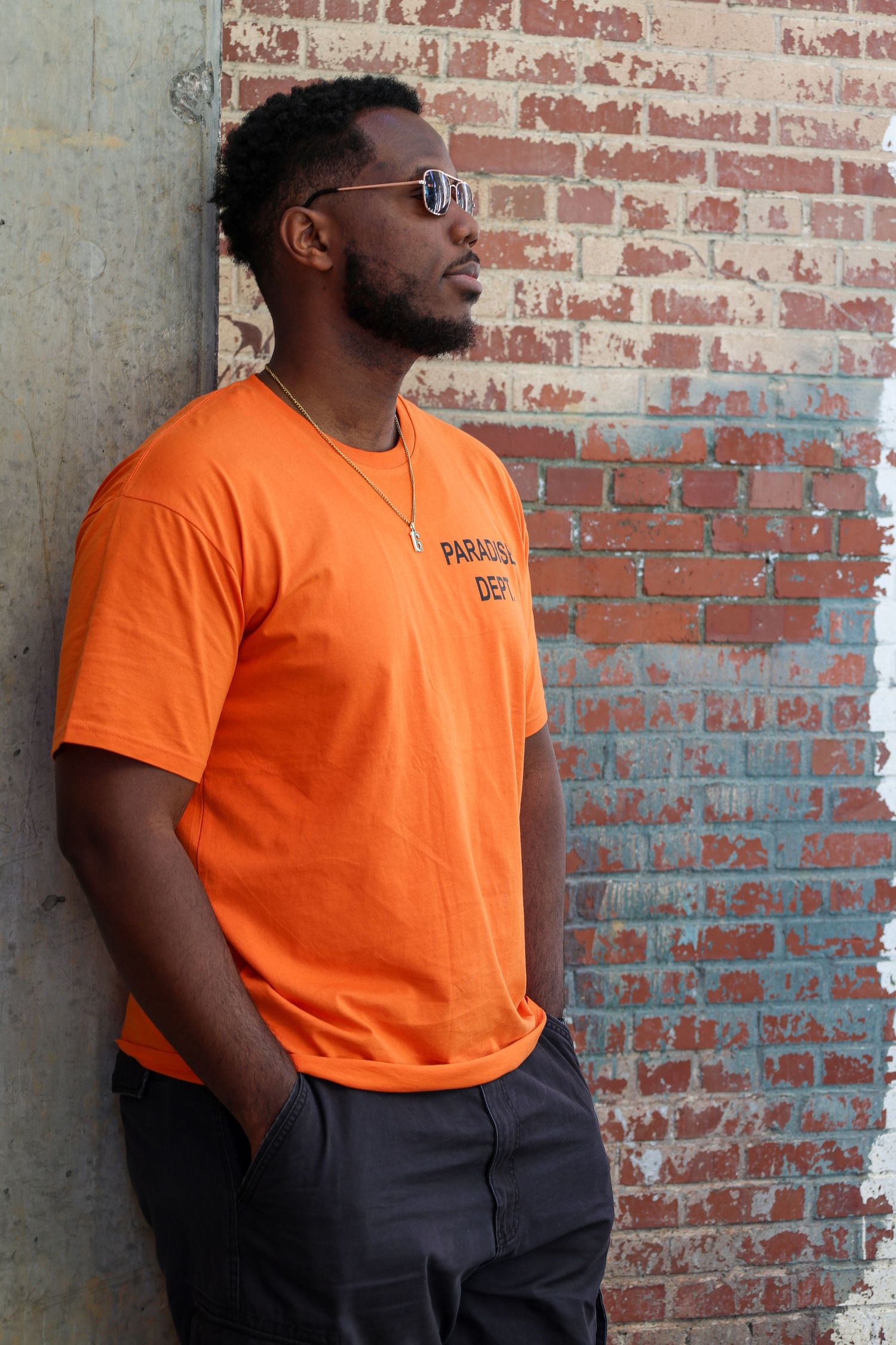 Man in orange graphic tee and sunglasses leaning against brick wall outdoors.