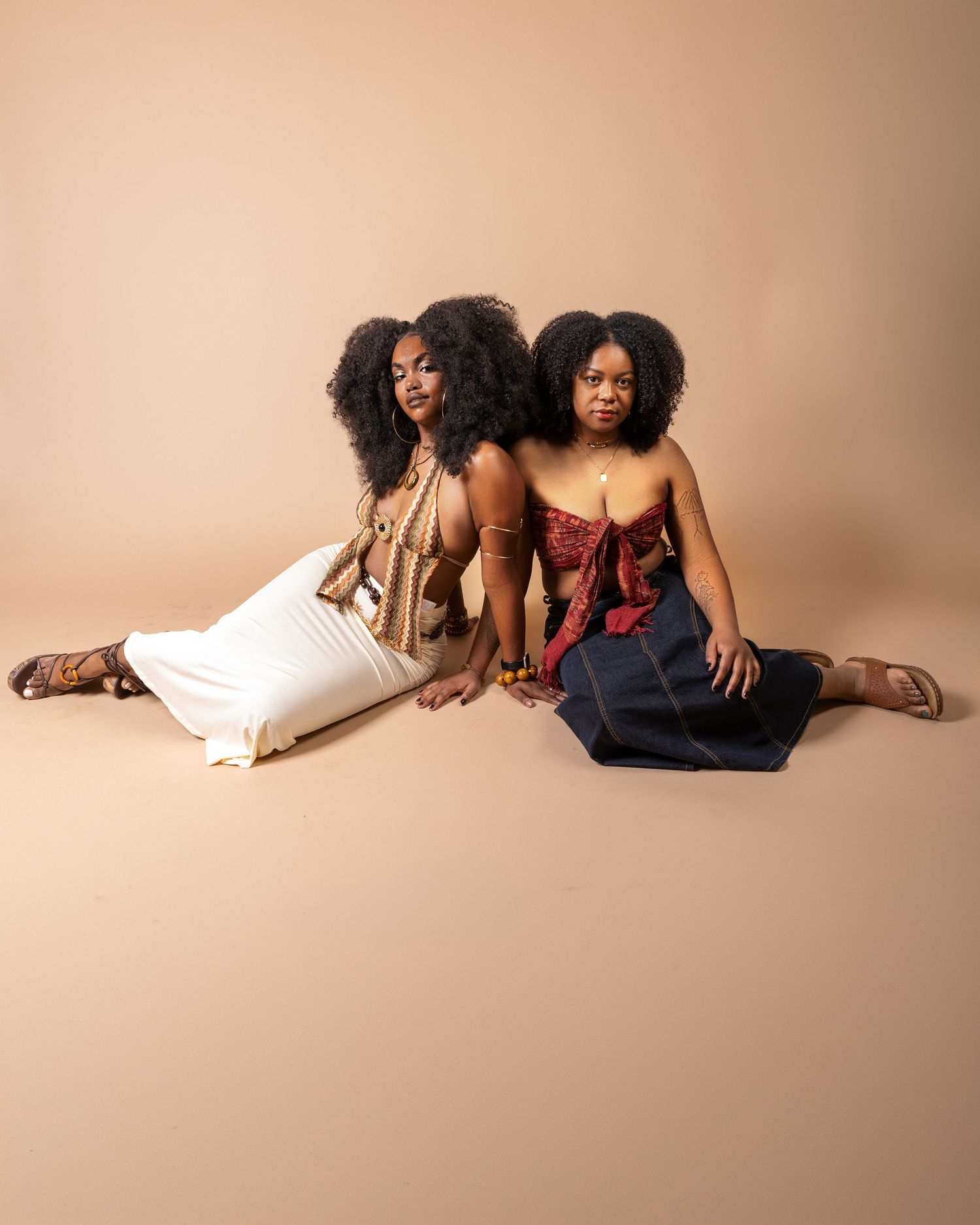 Two Black women with natural hair sit back-to-back on floor, wearing stylish outfits against a beige background.