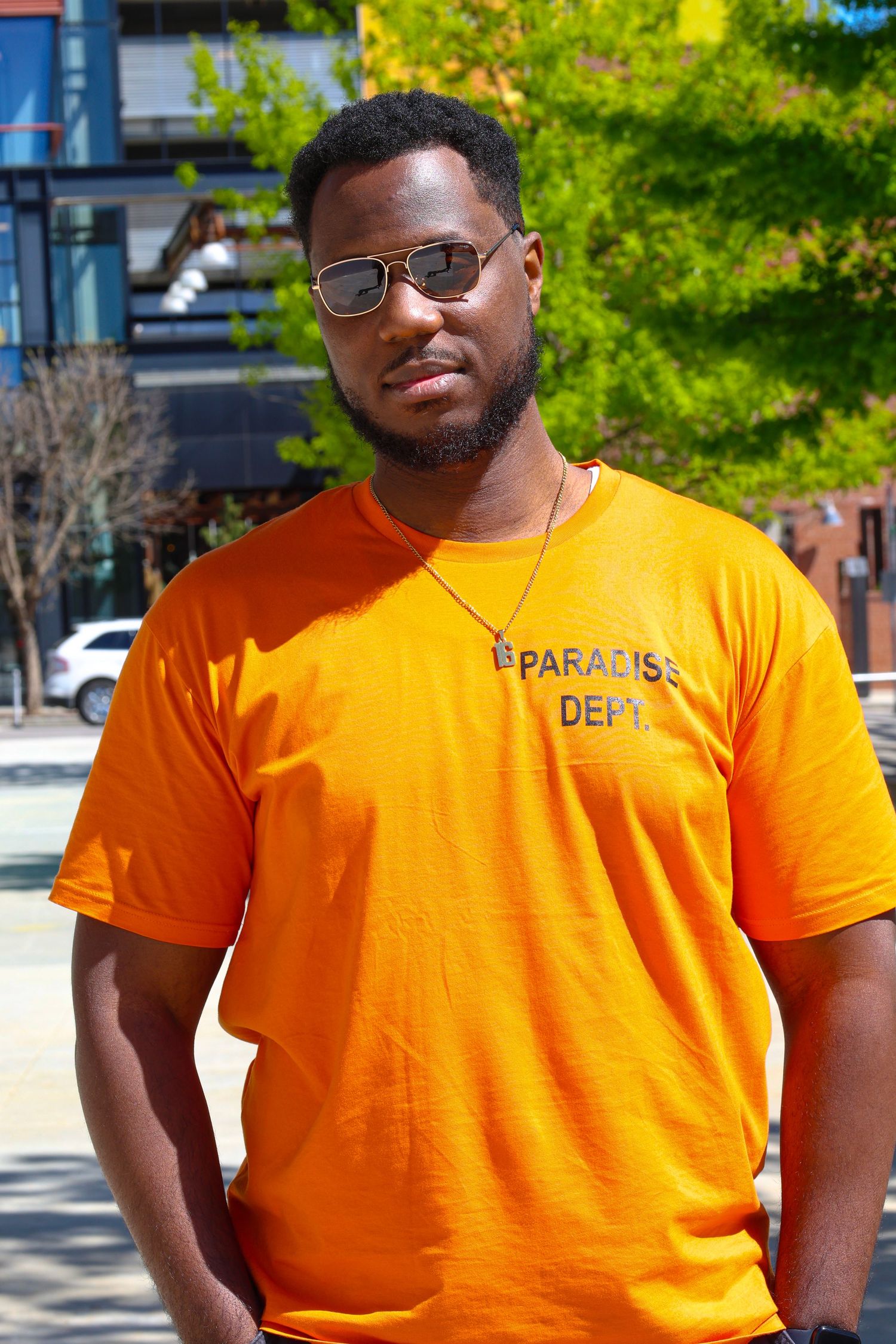 Man in orange Paradise Dept t-shirt and aviator sunglasses posing outdoors in urban setting.