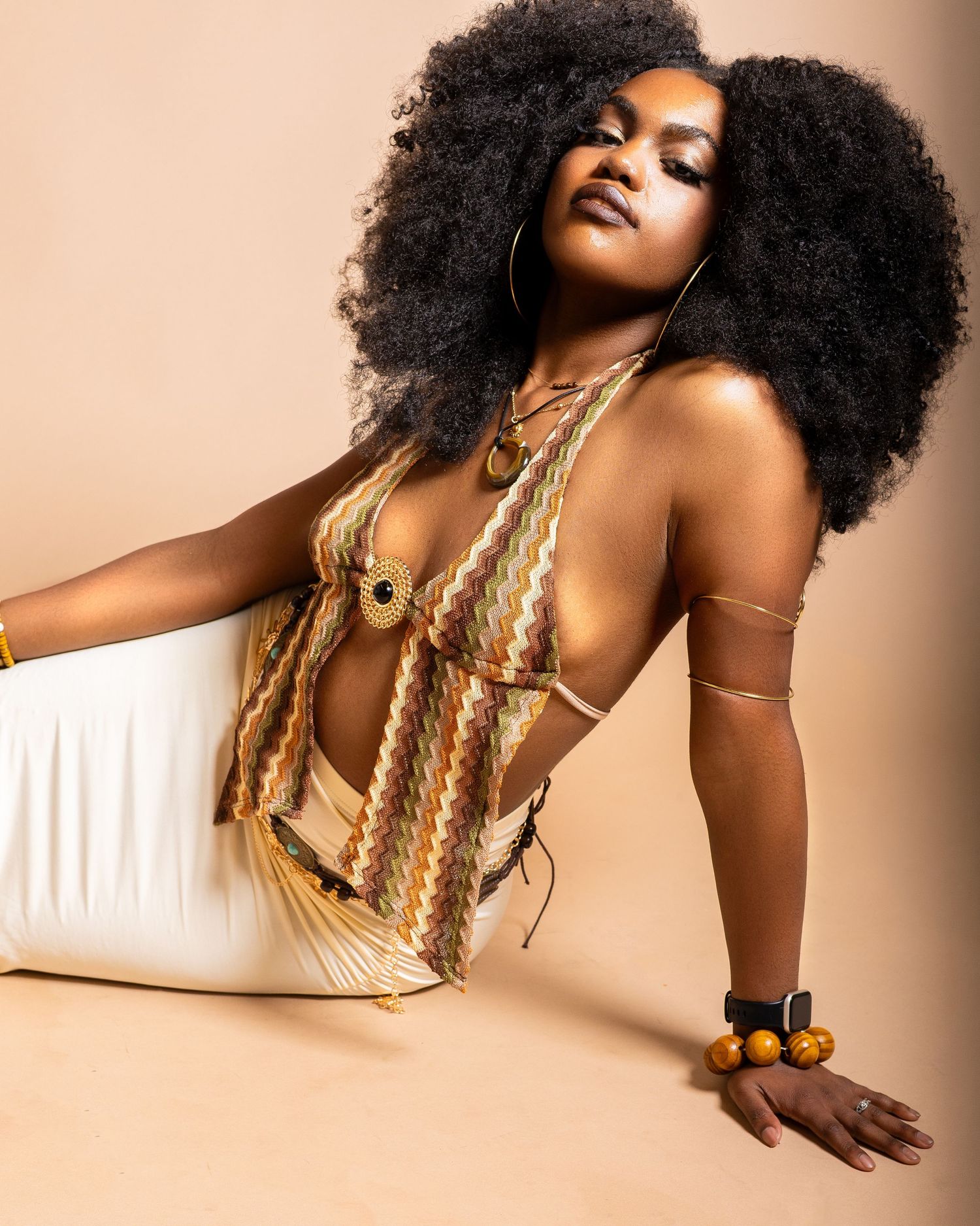 Woman with large afro in retro zigzag halter top, gold jewelry, and white pants poses confidently in studio.