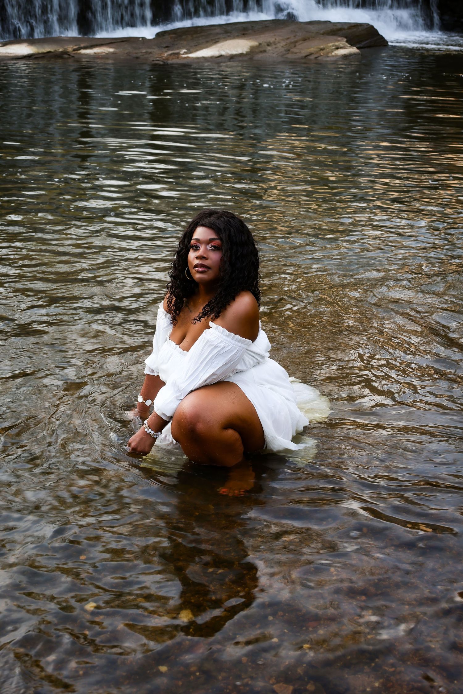 A woman in a white off-shoulder dress crouches gracefully in a shallow river with a waterfall in the background.