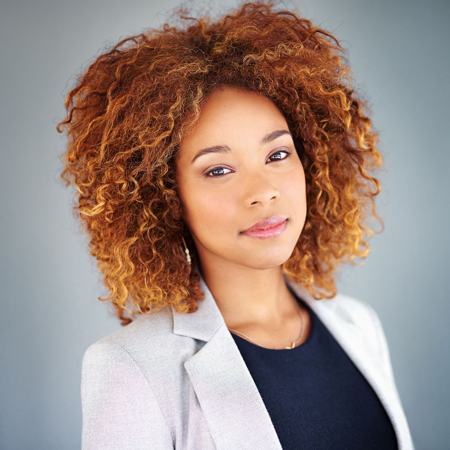Professional headshot of a woman with voluminous curly auburn hair wearing a light gray blazer.