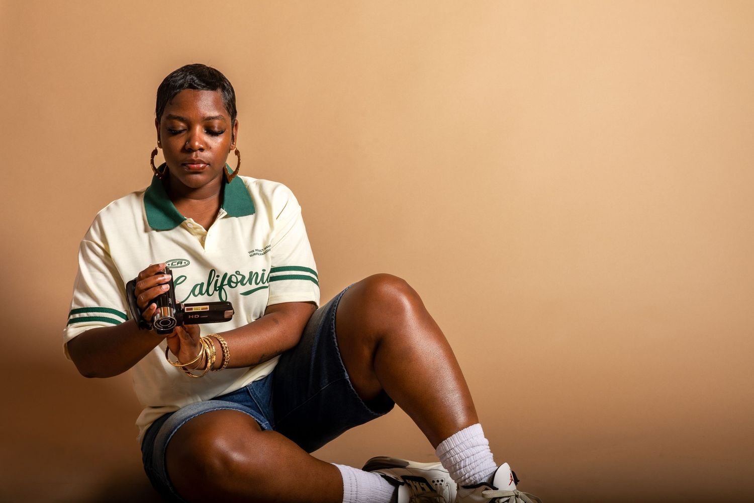 Woman in California jersey and denim shorts sitting casually against a tan background, looking at her phone.