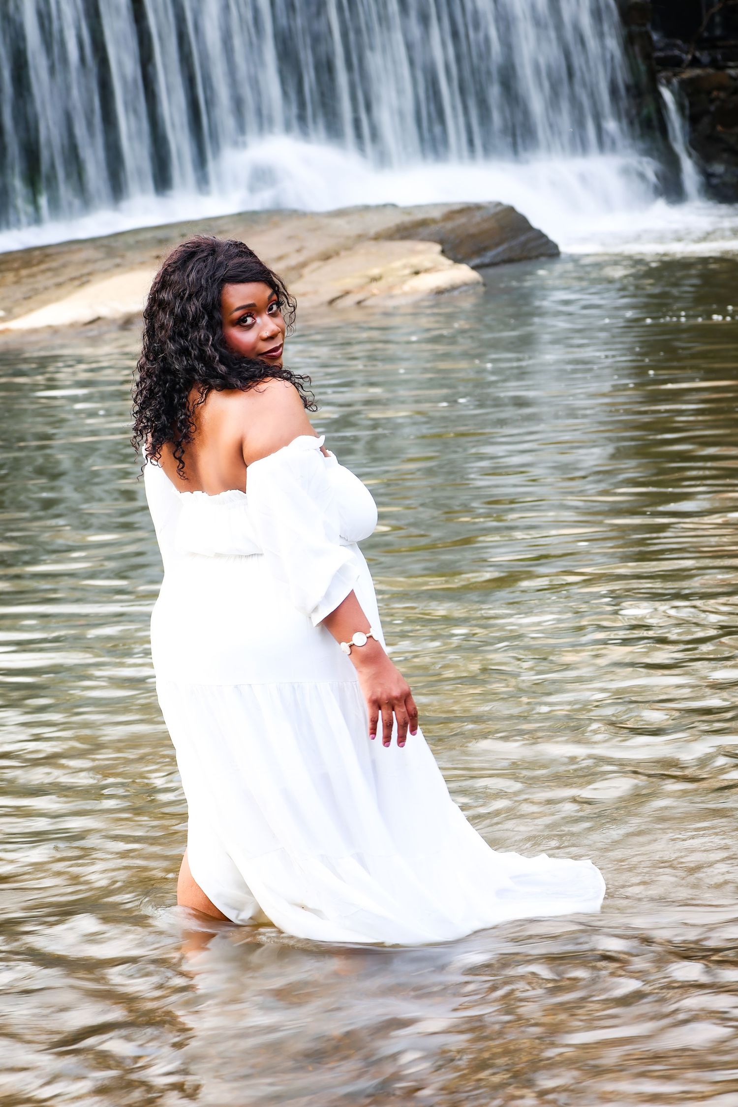 Woman in white off-shoulder dress standing in shallow water before a waterfall, looking back over her shoulder.