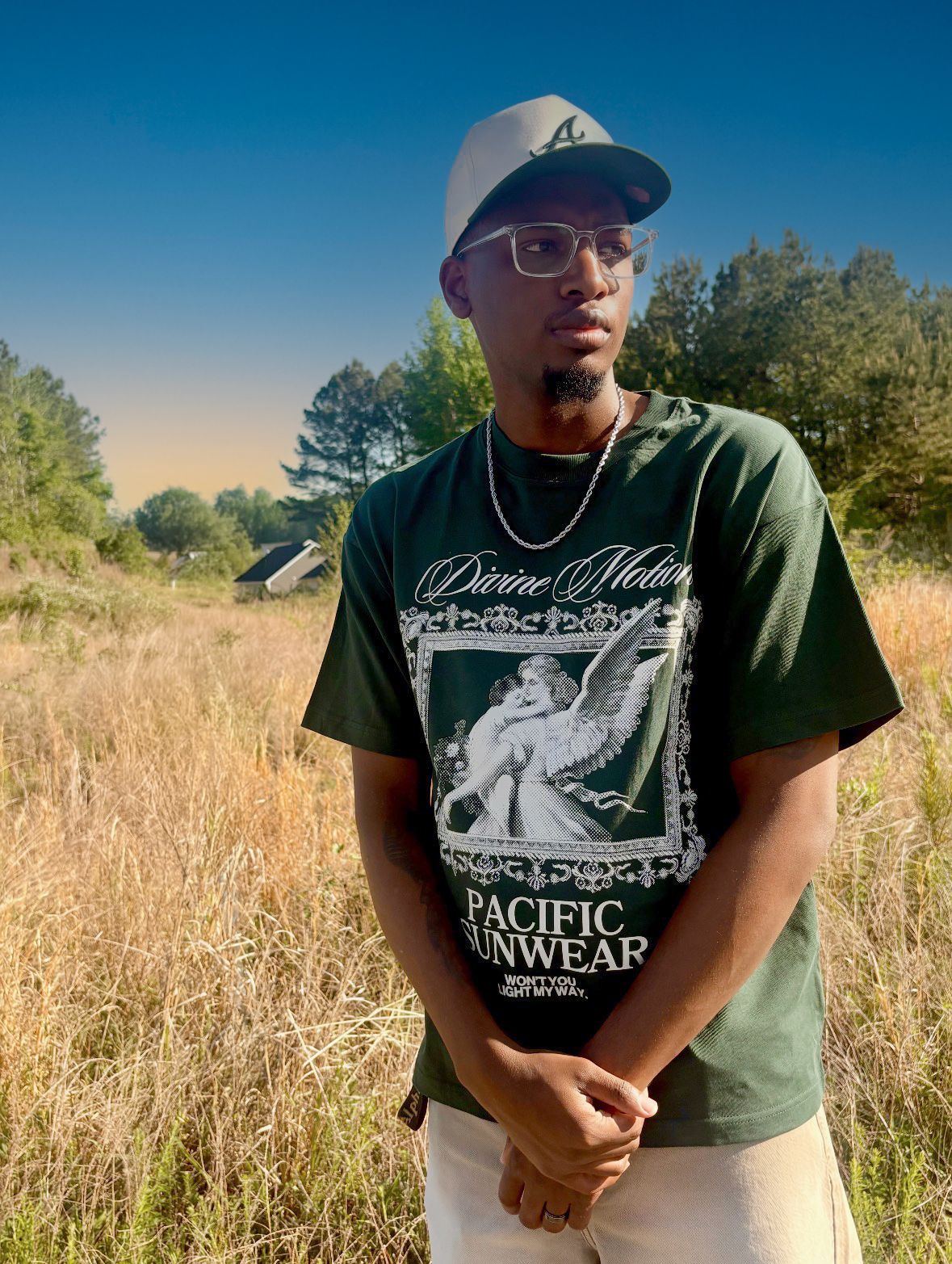 Young man in green Pacific Sunwear graphic tee and white cap standing in a golden field with trees and blue sky.