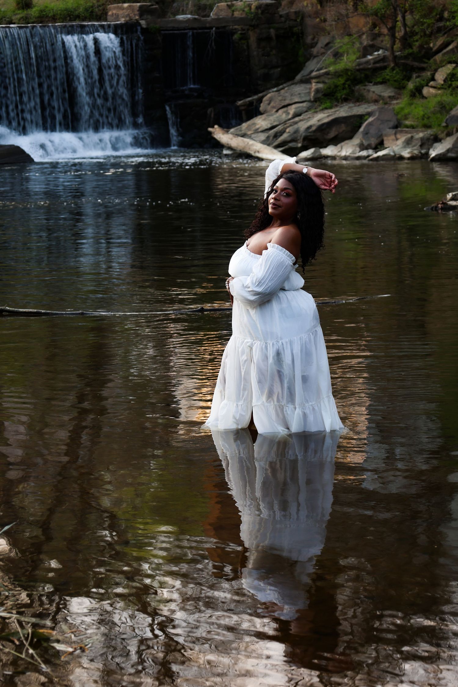 A woman in a white off-shoulder dress stands in a shallow river with a waterfall and rocky landscape behind her.