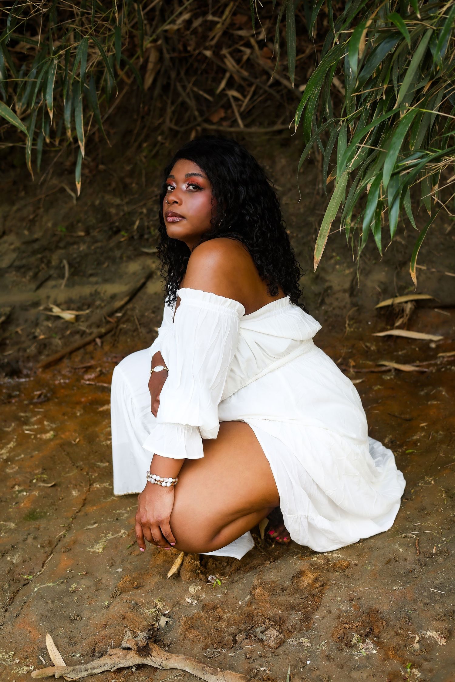 A Black woman in a white off-shoulder dress crouches gracefully on rocky ground surrounded by lush green foliage.