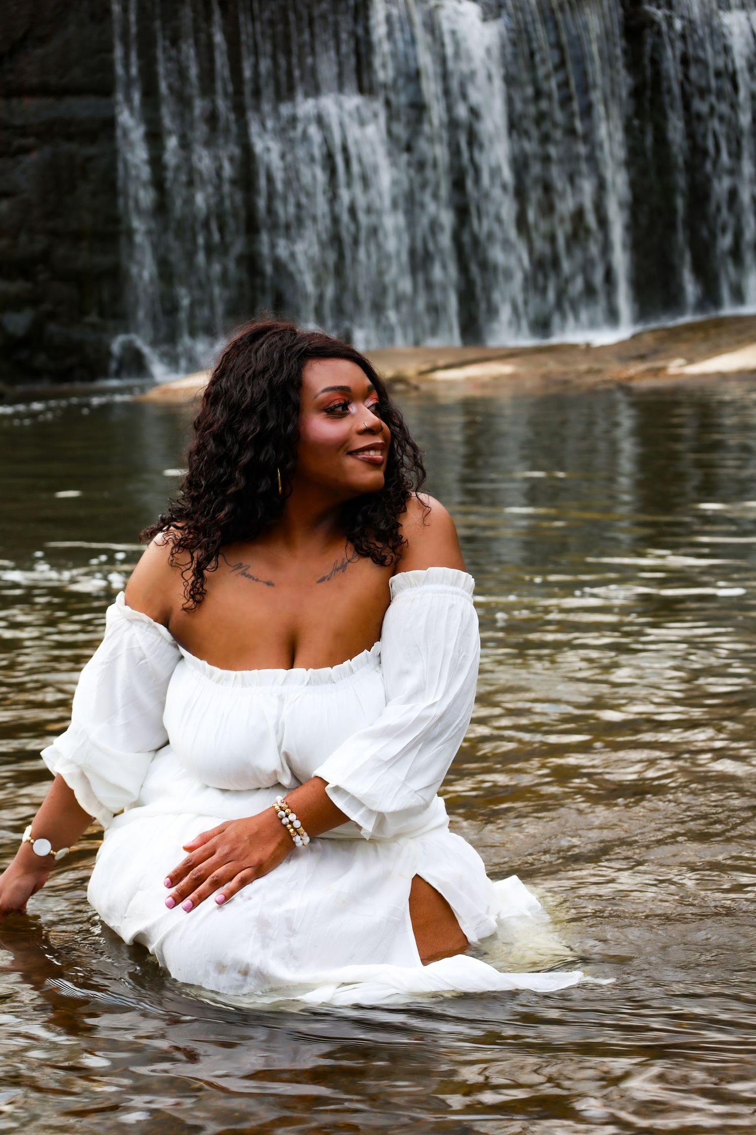 Woman in white off-shoulder dress sitting in water before a scenic waterfall, looking serene.