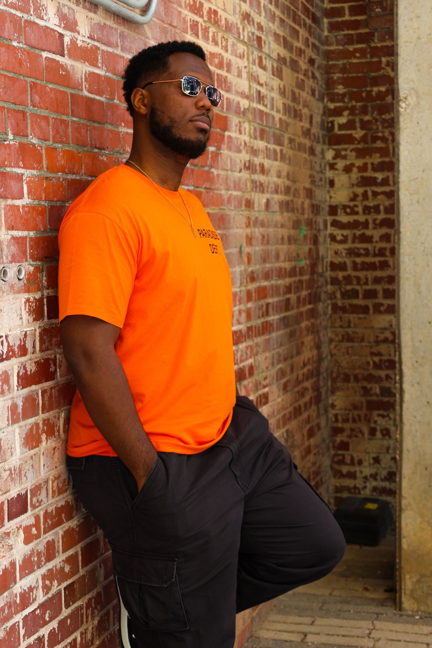 Man in orange Paradise Dept shirt leaning against red brick wall with one leg raised.