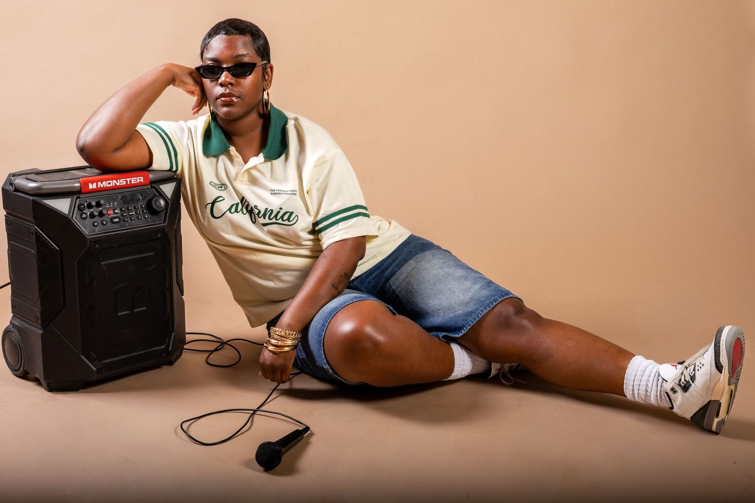 Man in sunglasses and denim shorts sits beside a portable speaker, holding a microphone on a beige background.