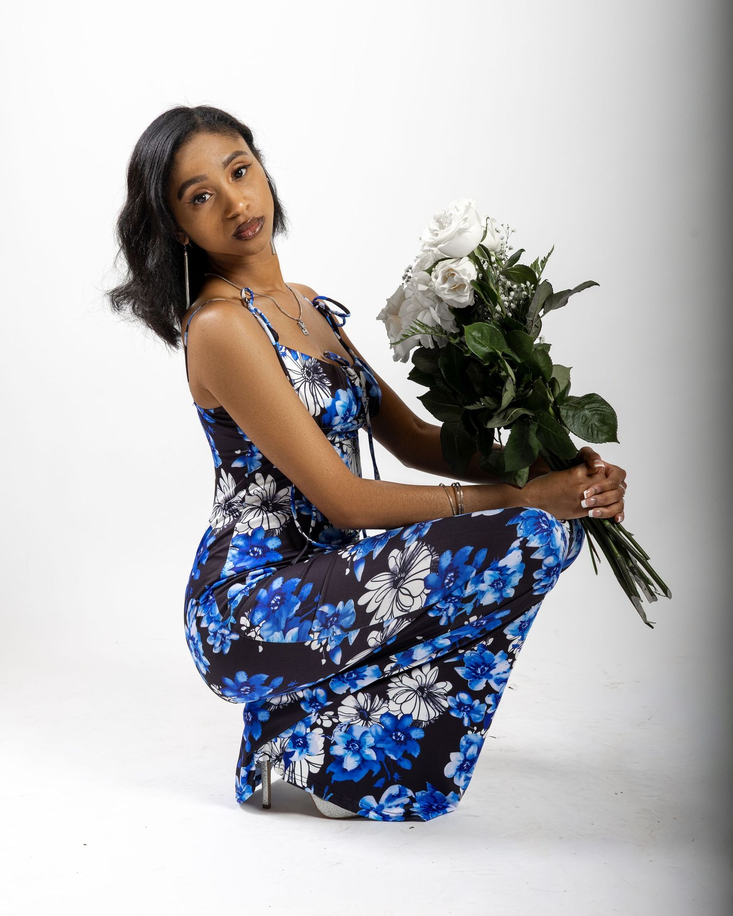 Woman in blue floral maxi dress kneeling while holding a bouquet of white roses against a white background.