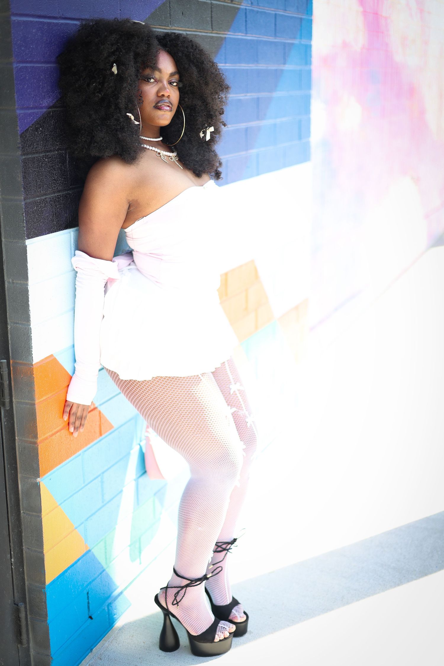 A stylish Black woman with voluminous afro hair poses in a white off-shoulder outfit with fishnet tights and black platform heels.