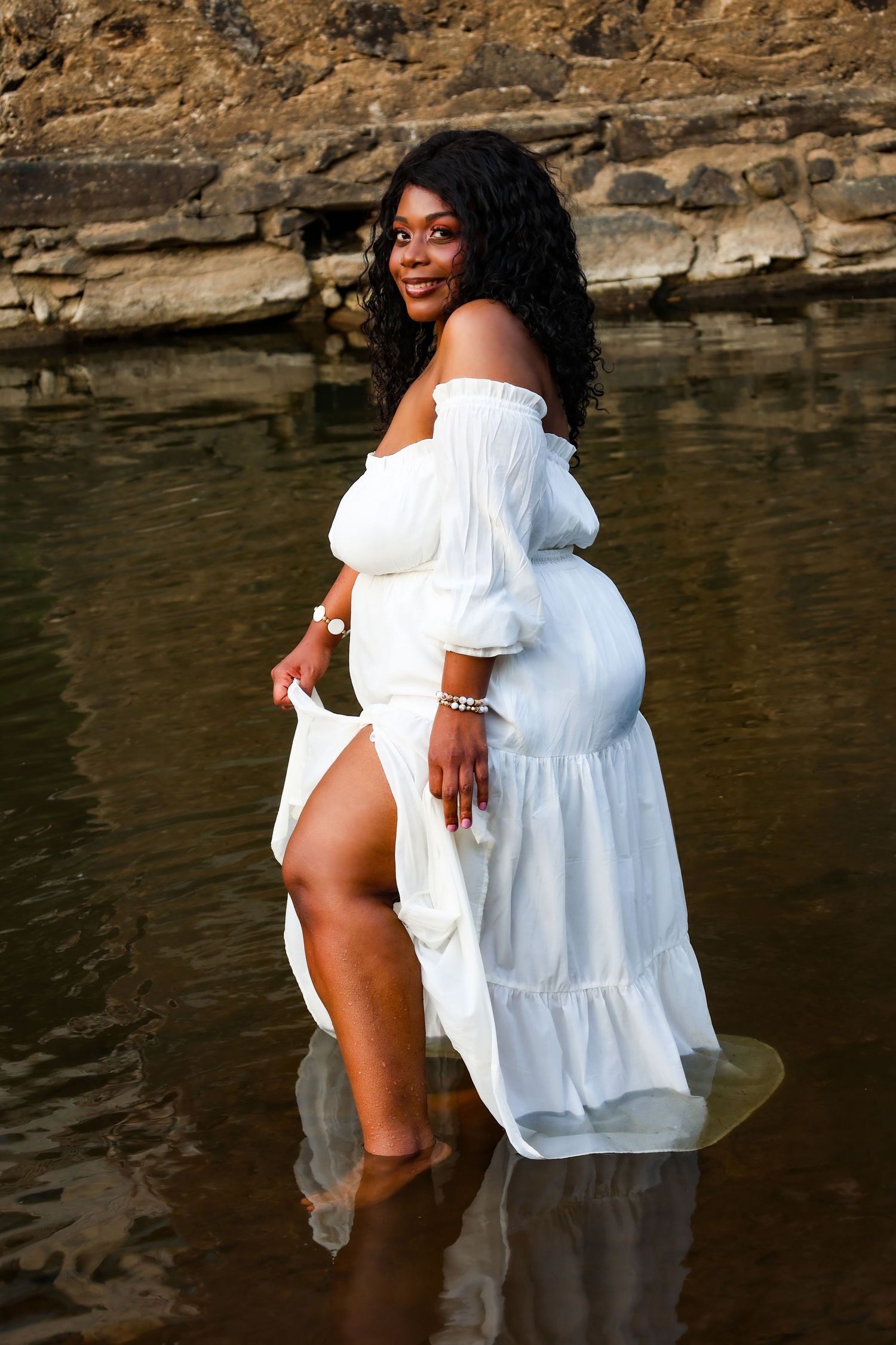 Pregnant woman in white off-shoulder dress standing in shallow water, smiling, with rocky background.
