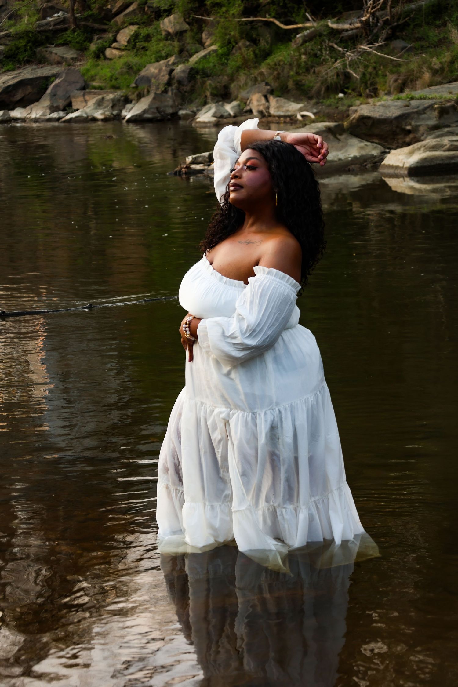 Woman in white off-shoulder dress standing in a rocky river, posing gracefully with hand in hair.