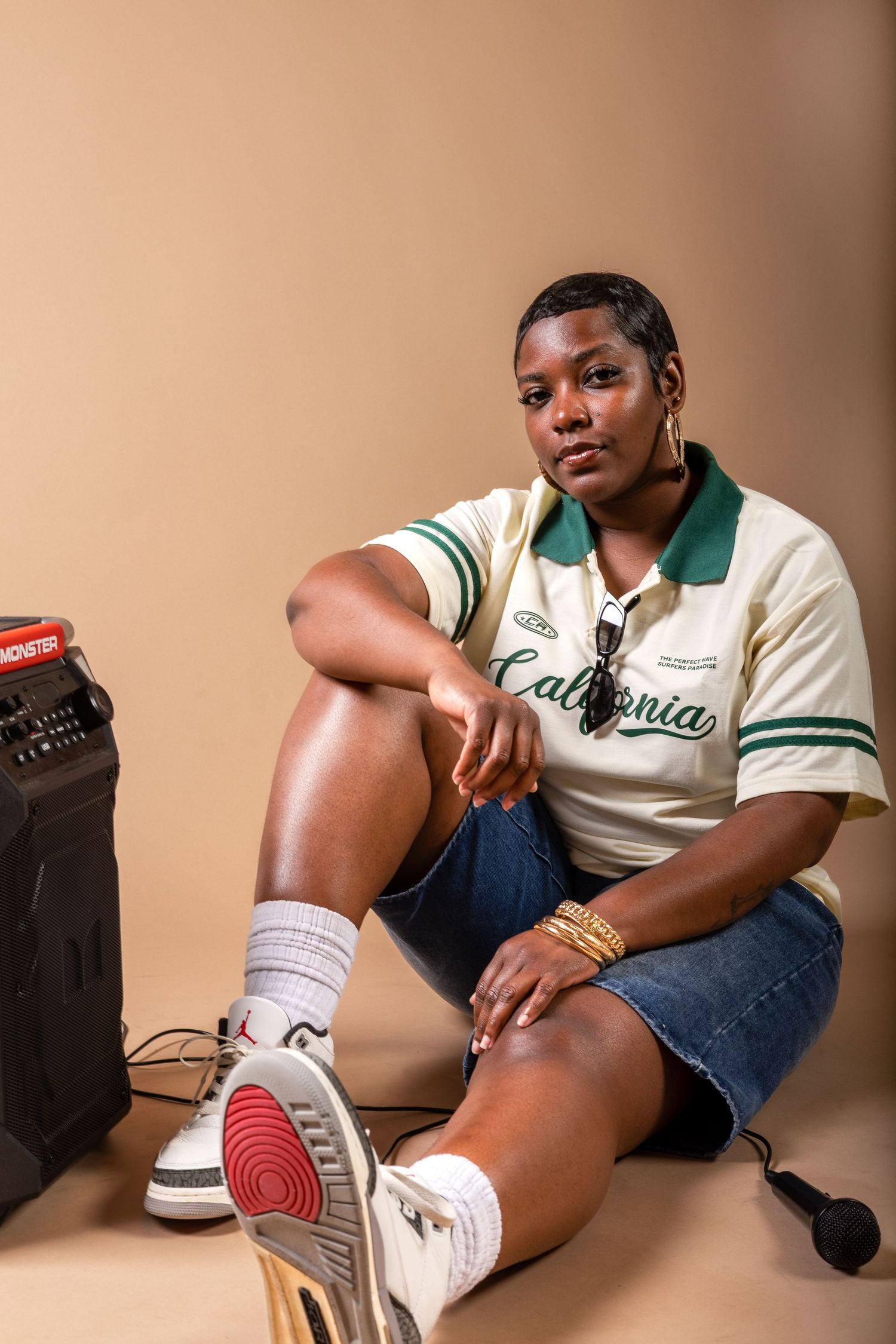 Young woman in California jersey and denim shorts sits beside a boombox with sneakers and gold jewelry.