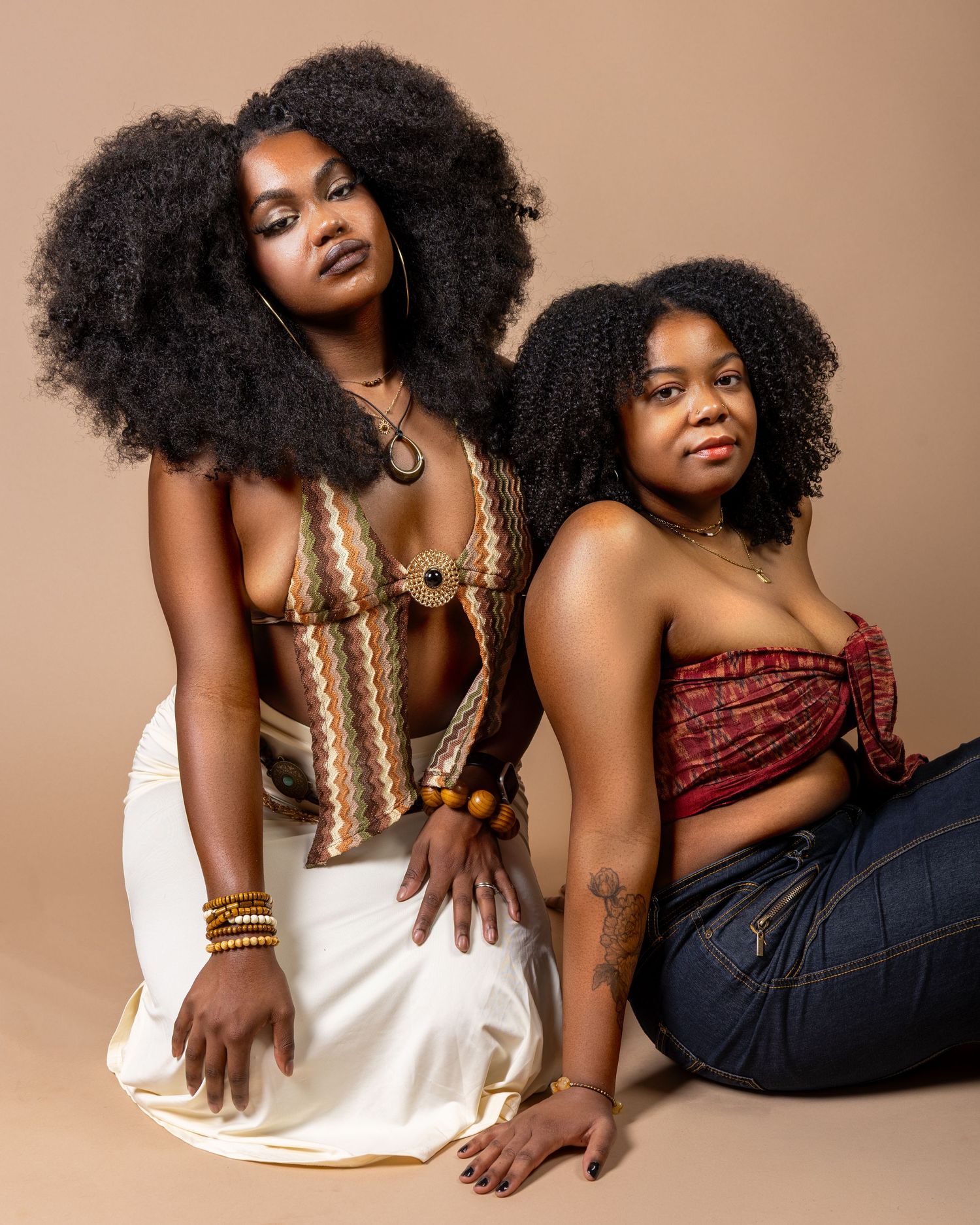 Two Black women with natural afro hair pose confidently in bohemian crop tops and jeans against a beige backdrop.