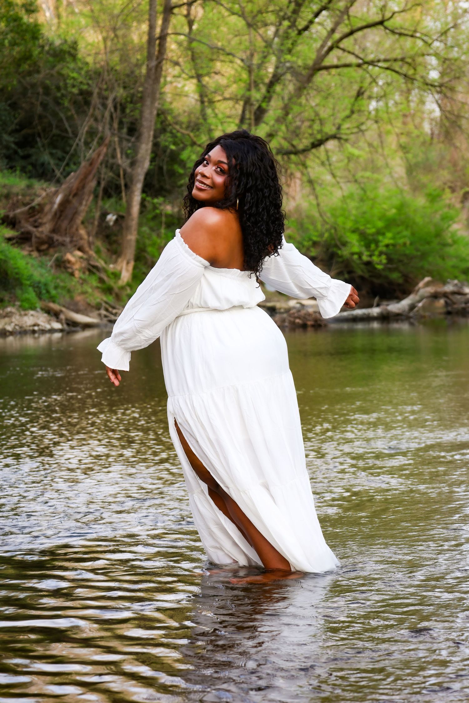A smiling Black woman in a white off-shoulder gown wades through a serene river surrounded by lush green trees.
