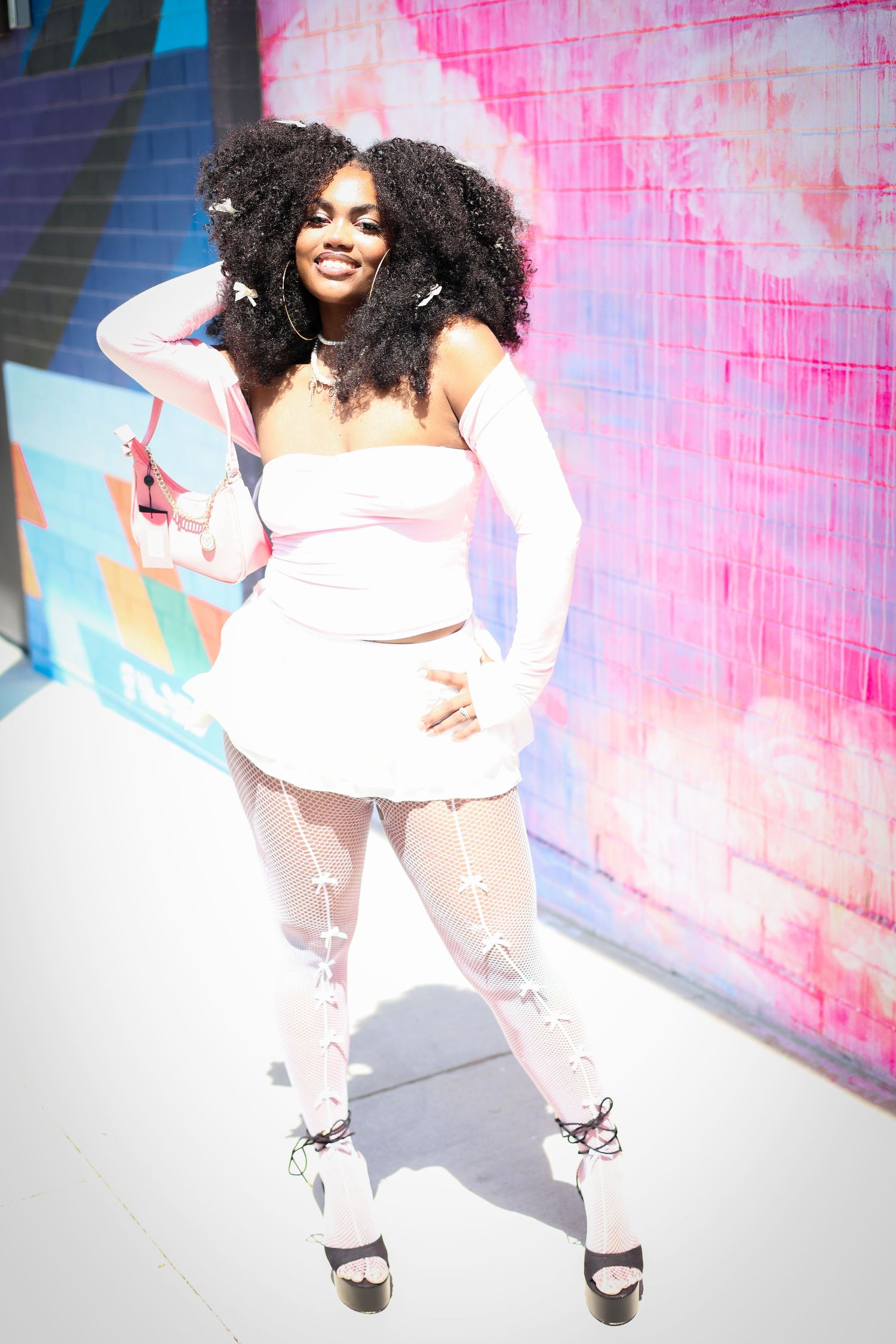 A Black woman in a white crop top, mini skirt, fishnet tights, and long gloves poses confidently outdoors.