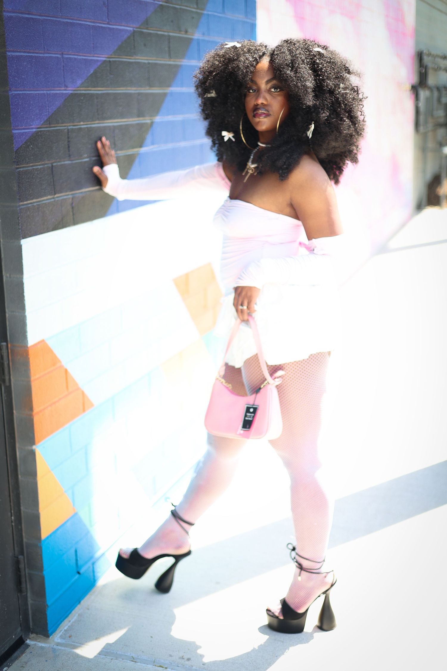 Black woman with afro in pink outfit and black platform heels poses against colorful mural wall holding pink bag.