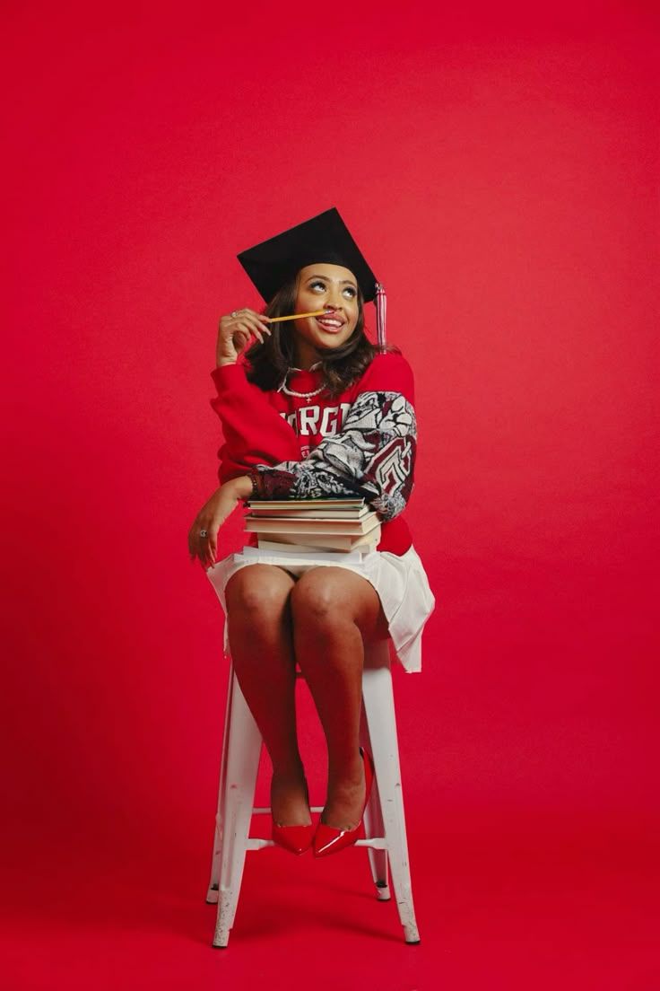 Smiling woman in graduation cap and red college sweatshirt sitting on stool with stacked books.