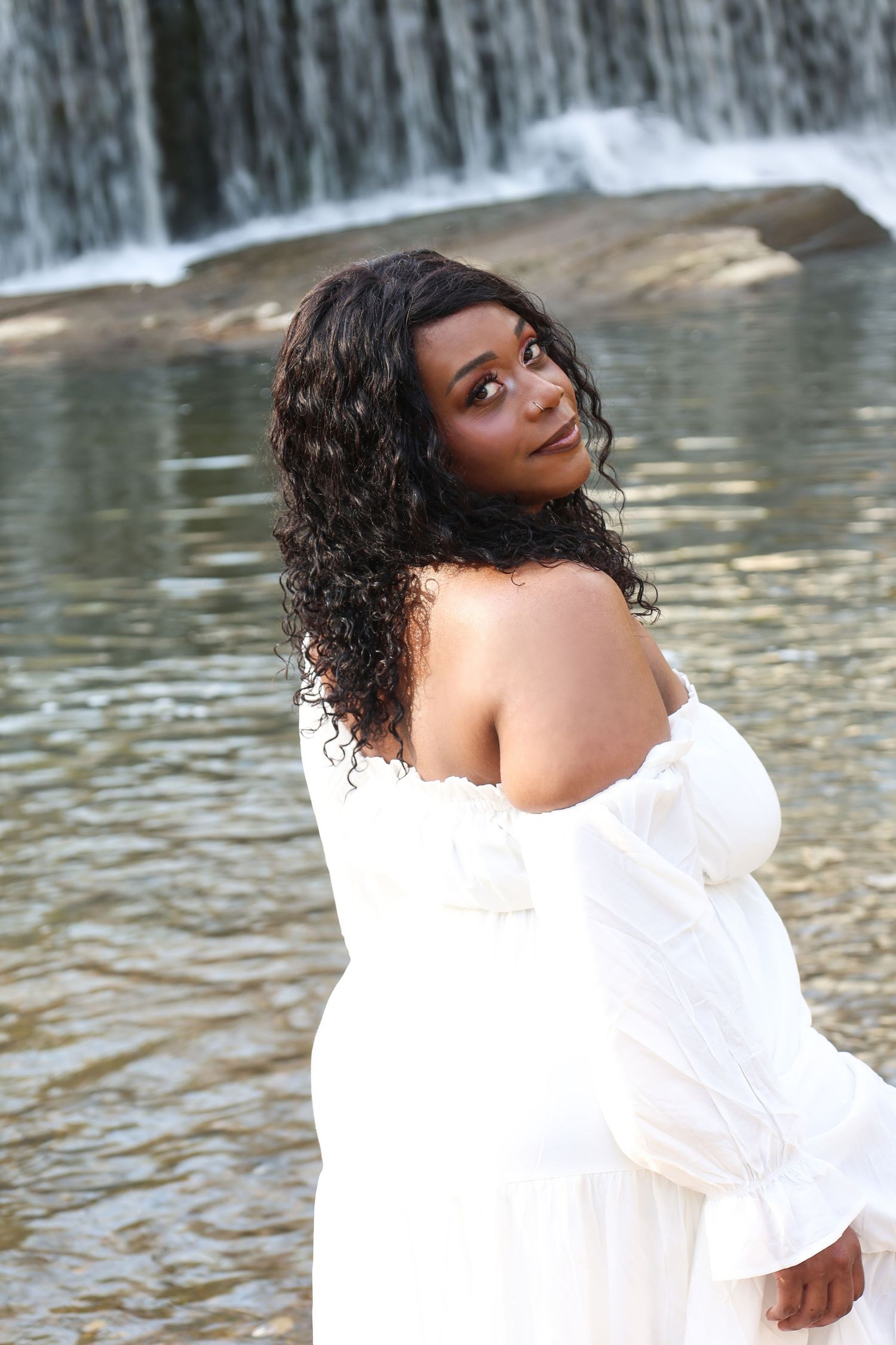 A woman in a white off-shoulder dress poses gracefully in front of a scenic waterfall with curly dark hair.