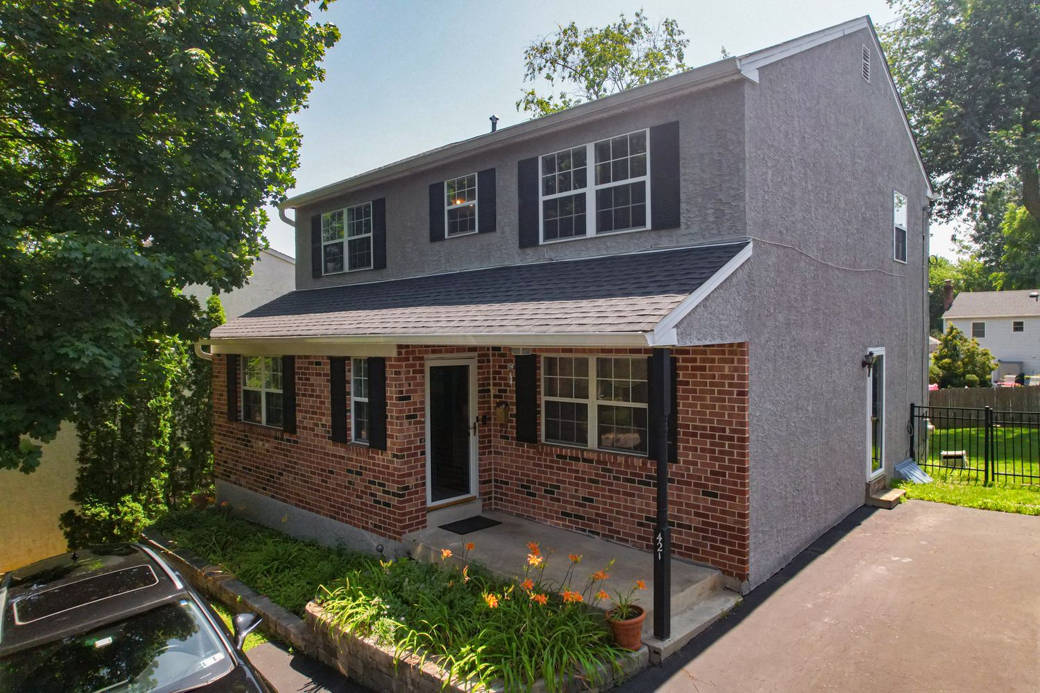 Two-story colonial home with brick facade, gray siding, black shutters, covered entry, and lush green landscaping.
