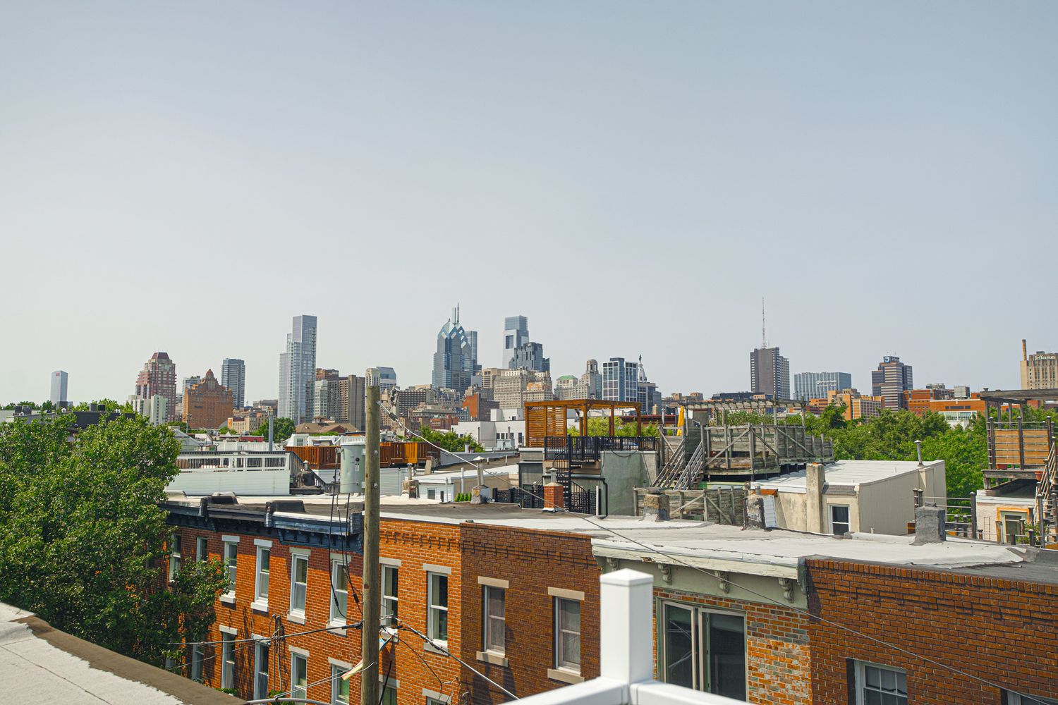 Rooftop view of Philadelphia city skyline with brick row houses, green trees, and skyscrapers under a hazy sky.