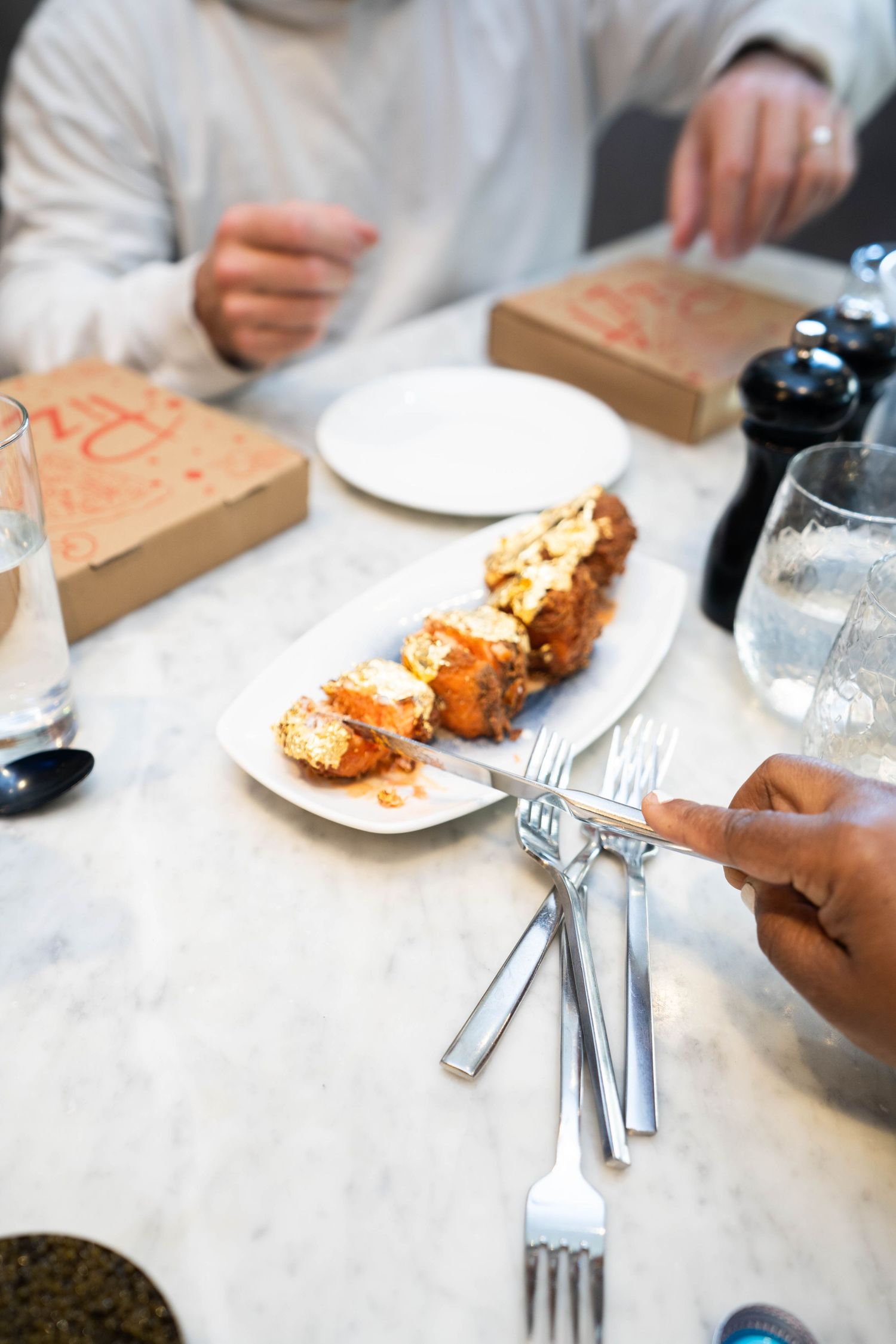 People dining at a marble table with appetizers on a white plate, silverware, and small cardboard boxes at a restaurant.