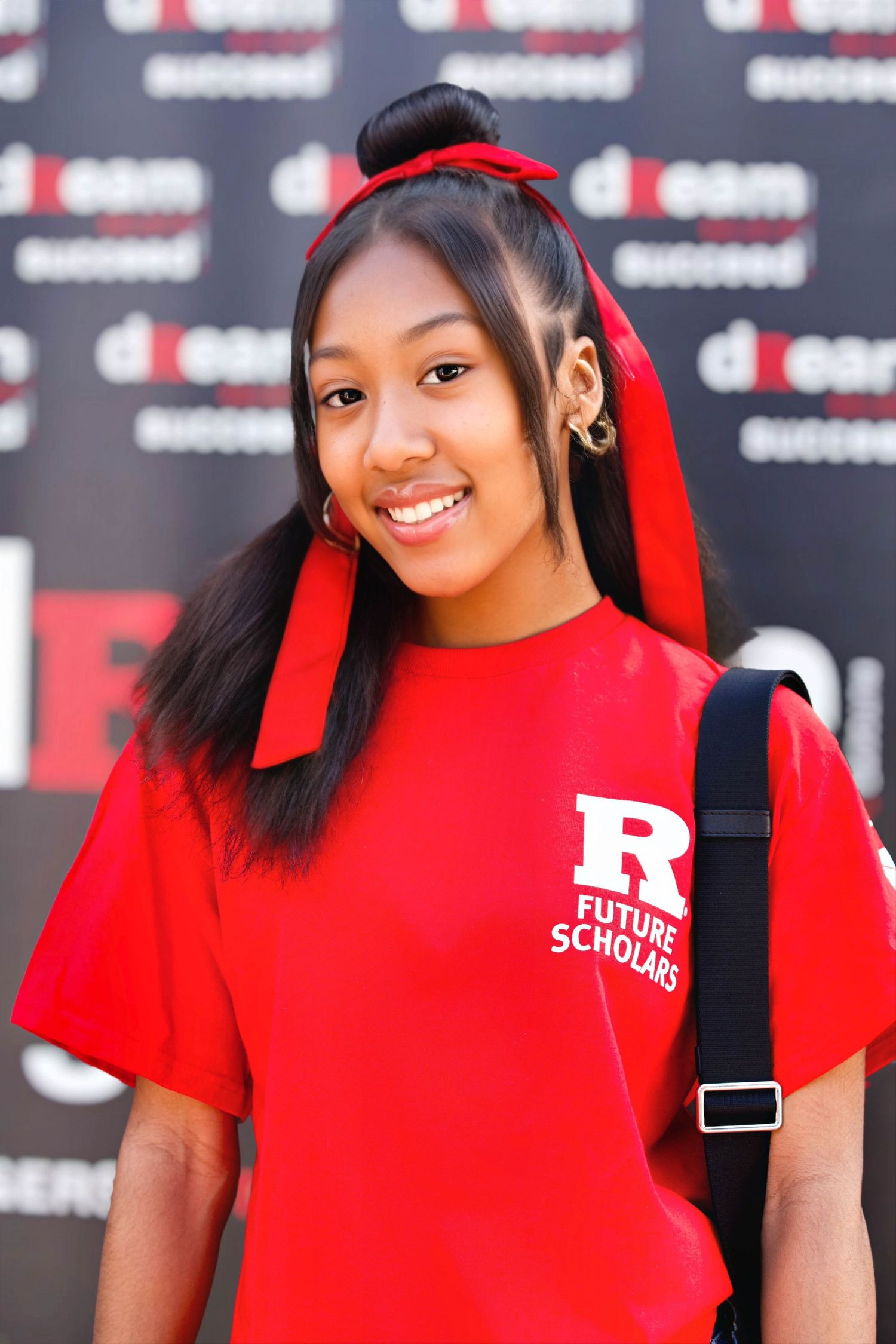 Smiling young woman in red Rutgers Future Scholars t-shirt with red hair bow, standing in front of a dream succeed banner.