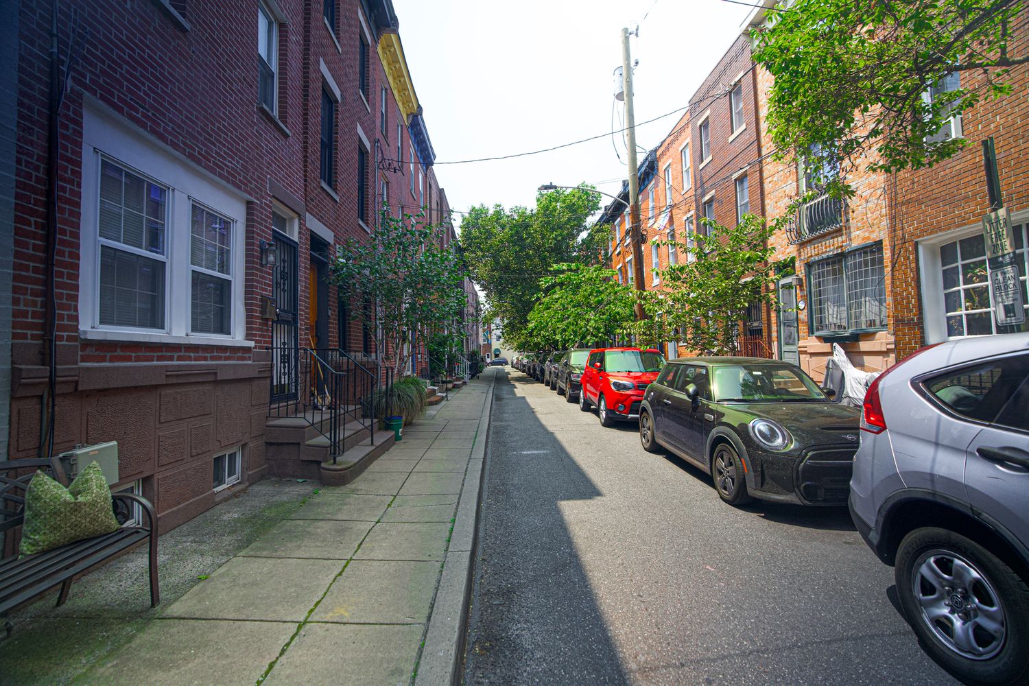 Narrow residential street lined with red brick rowhouses and parked cars in an urban Philadelphia neighborhood.