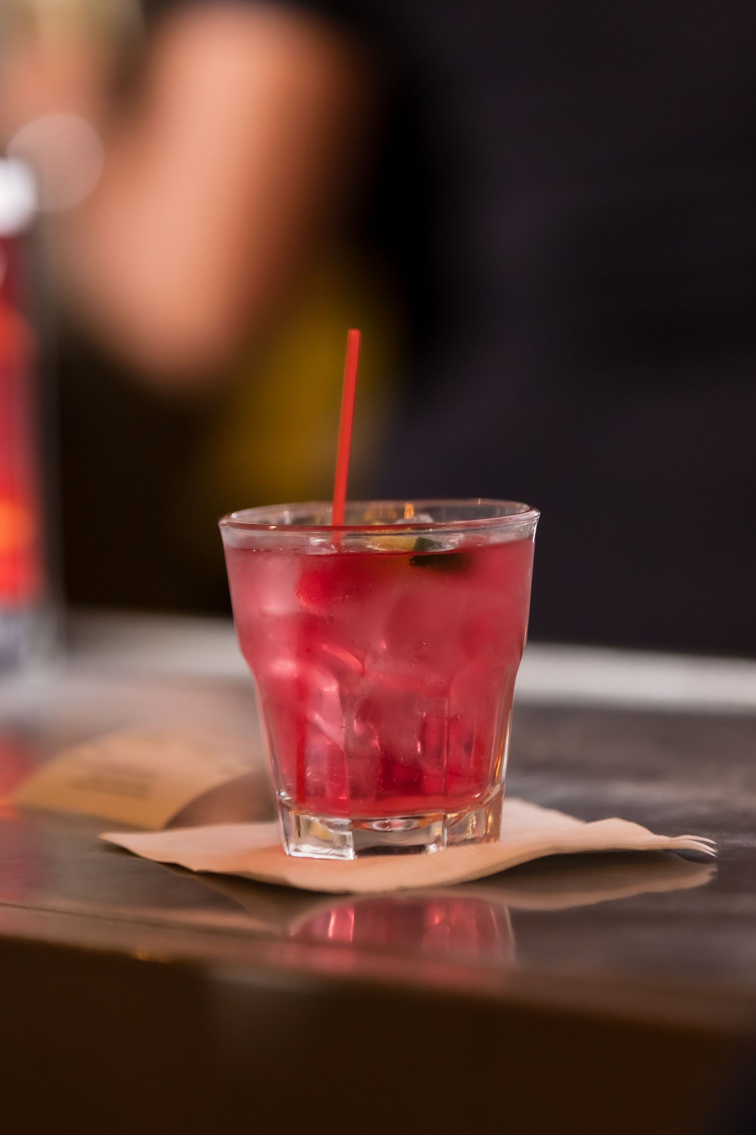 A red cocktail with ice and a straw served on a napkin at a bar counter.