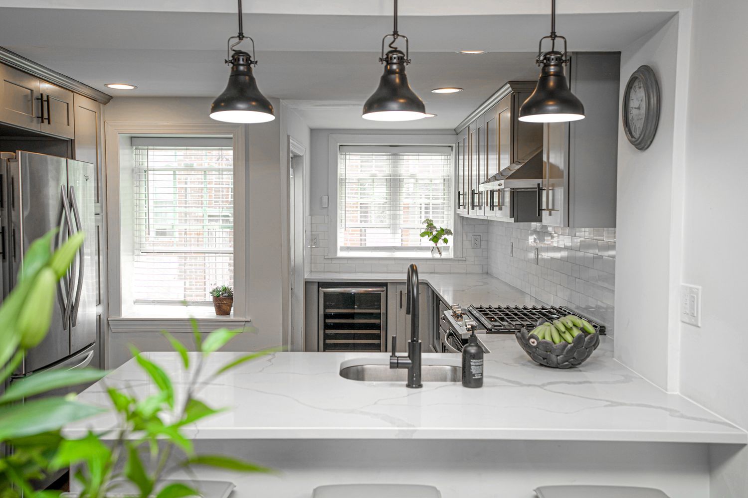 Modern white kitchen with marble countertops, black pendant lights, stainless steel appliances, and large windows.