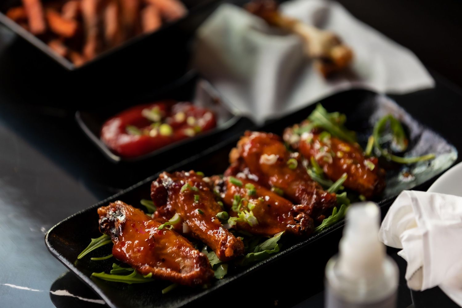 Glazed chicken wings garnished with green onions served in a black dish with dipping sauce.