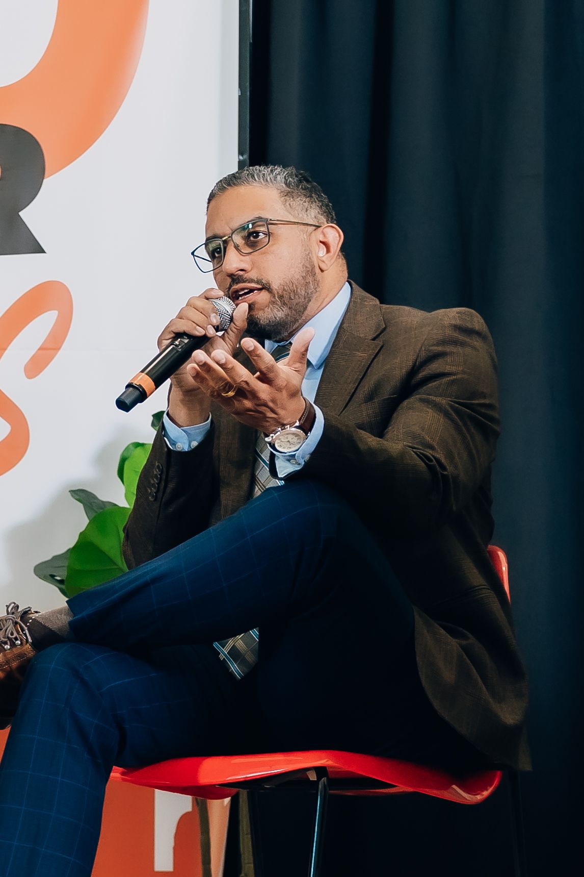 Man in brown blazer speaks into microphone while seated on red stool at a panel discussion event.