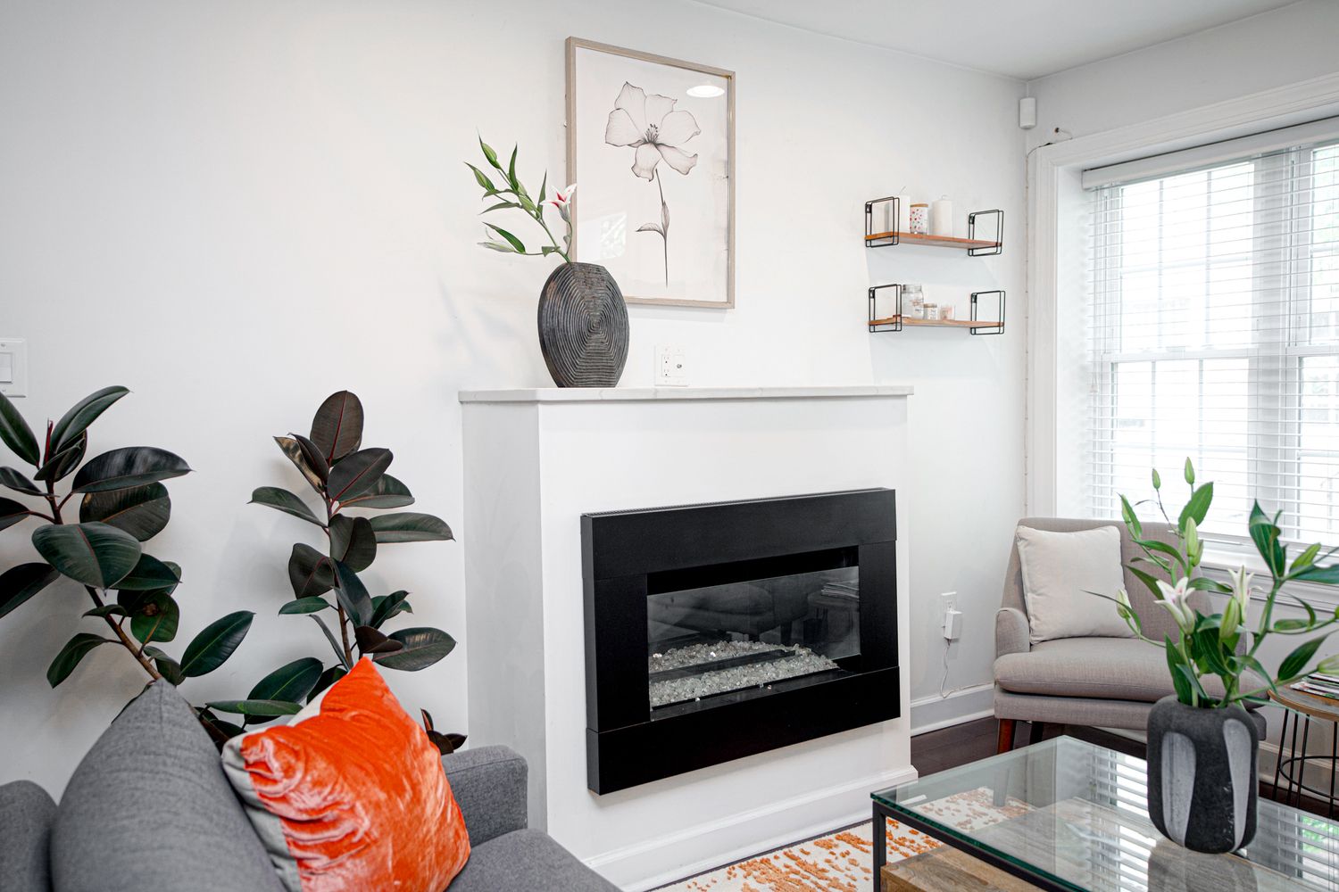 Modern living room with white fireplace, decorative mantel, gray sofa, orange pillow, and rubber plant near window.